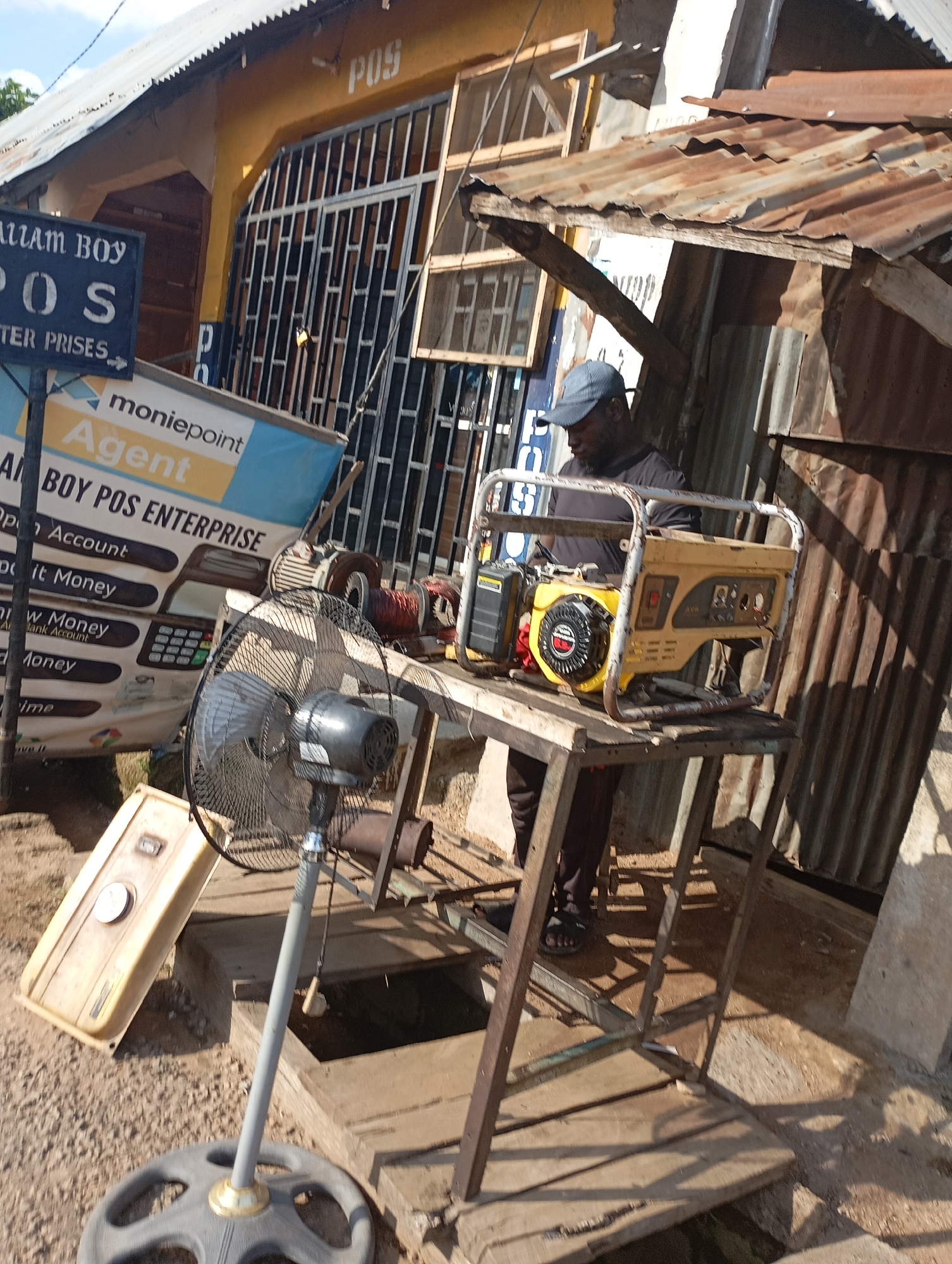 In Shendam, Nigeria, a man wearing a grey baseball cap and a dark shirt stands beside a yellow generator on a metal stand. In front of the generator, a black oscillating fan on a grey pole faces towards the viewer. To the left of the fan, a beige plastic container is placed on the ground. Behind the man and the generator is a structure with a corrugated metal roof and walls, and to the left, a building with a yellow facade is visible. A sign that reads "MONIEPOINT AGENT" is prominent on the left side of the image. The ground is a mix of gravel and concrete.