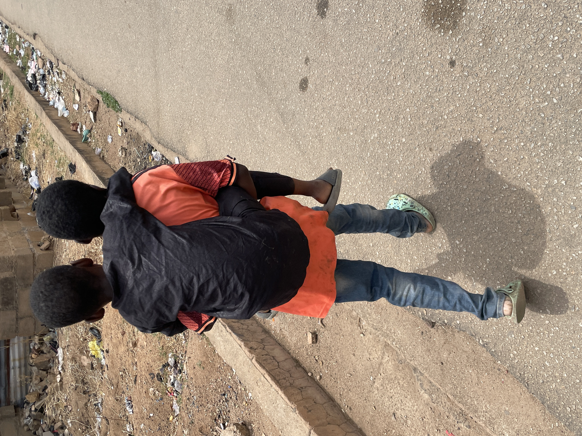 Two boys are on a sidewalk next to a street in Jos, Nigeria. One boy is giving the other a piggyback ride. The boy carrying has on blue jeans, olive colored shoes, a burnt orange shirt and black jacket. The boy being carried has on black shorts and a burnt orange shirt. There is a lot of trash along the side of the street.