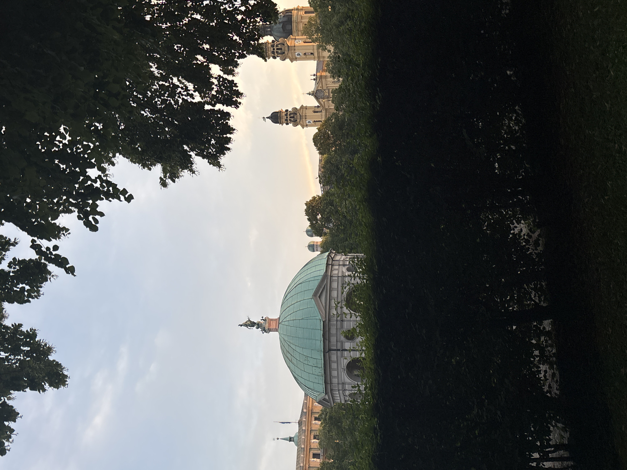 The image depicts the exterior of the Monopteros in the Englischer Garten, Munich, Germany. The Monopteros, a classical temple, is partially obscured by a dark green hedge in the foreground. The temple features a distinctive green dome topped with a statue. Behind the Monopteros, other structures are visible, including a building with two prominent towers, bathed in the warm glow of the setting sun. The sky above is a pale blue with scattered clouds, and the branches of trees with dark green leaves frame the top of the image. The overall scene is one of serene architecture and natural beauty at dusk.