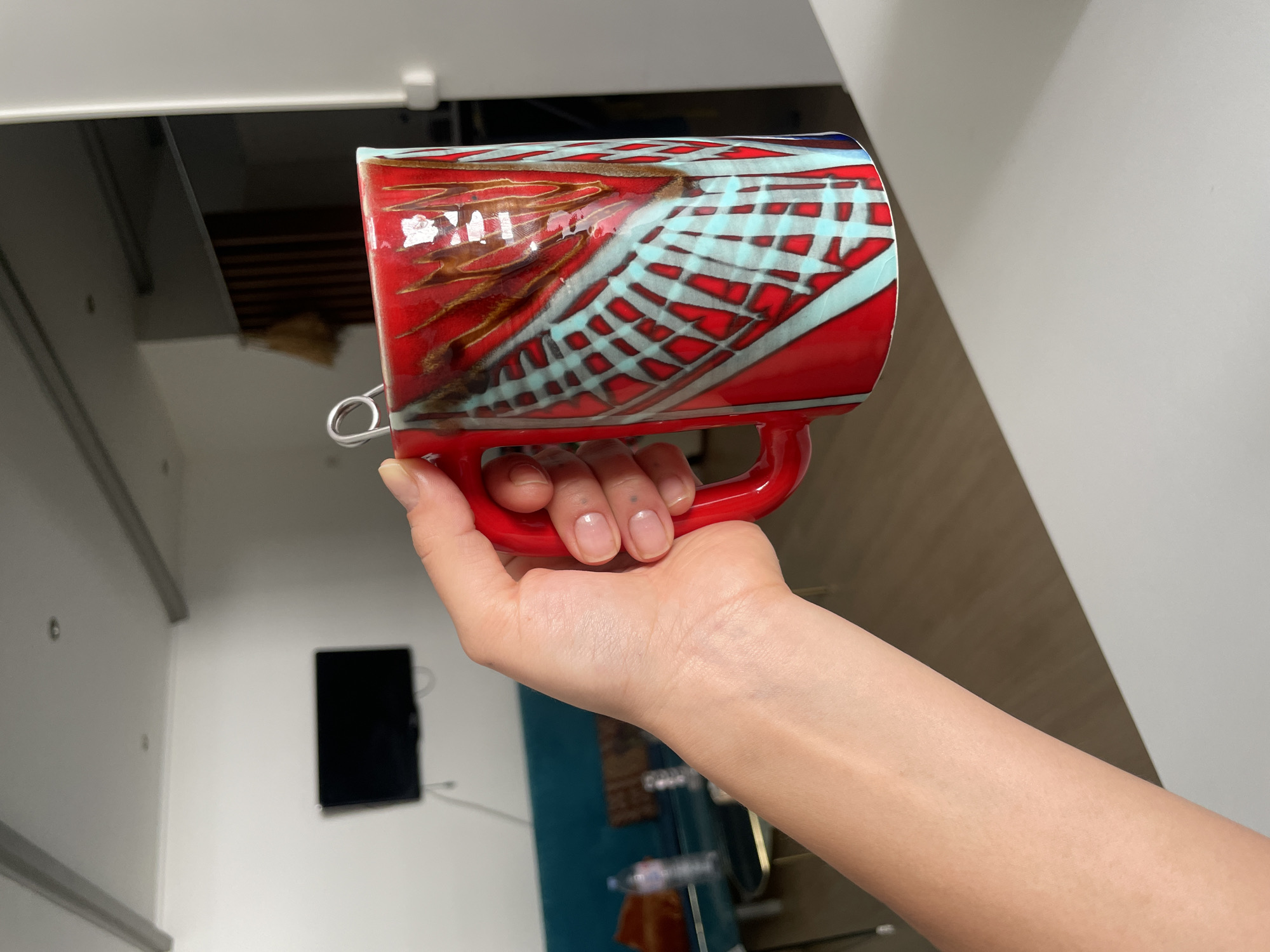 A person is holding a red mug with a unique, hand-painted design featuring red, brown, and blue colors in an abstract grid and swirl pattern. The mug has a red handle and a silver-colored metal ring attached near the top. The person's hand is visible, with bare fingernails. The background is out of focus but appears to be a domestic interior, possibly a kitchen or living space. There is a television mounted on the wall, along with cabinets. The mug appears to be in a modernly decorated apartment, but no specific landmarks of Paris are present to identify the exact location.