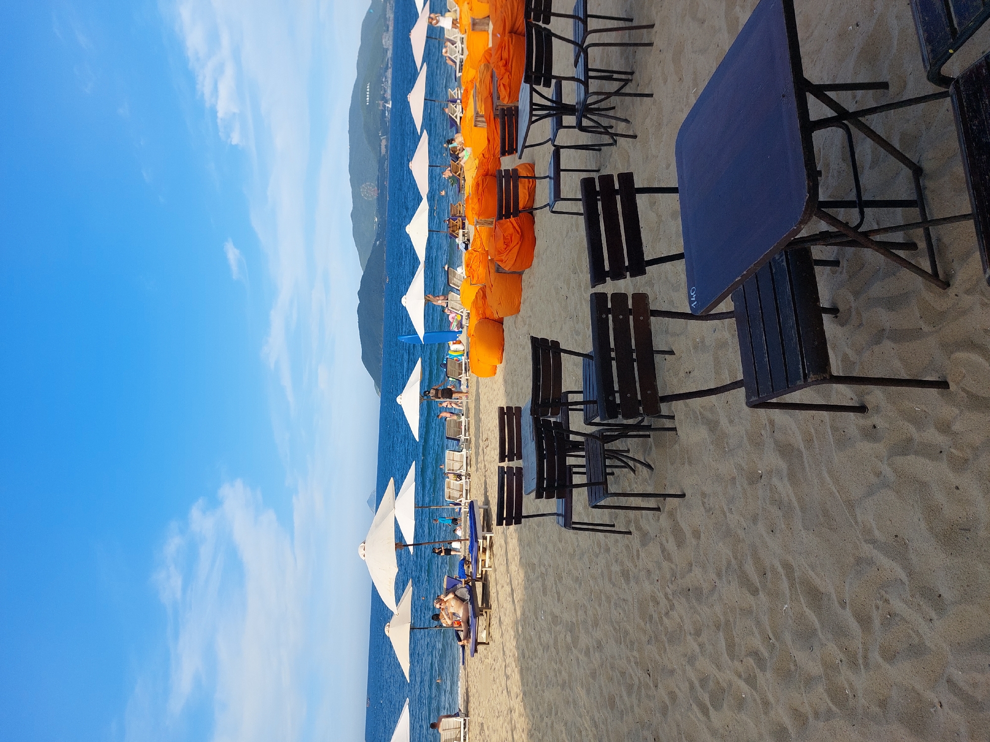 The image depicts a beach scene in Nha Trang Ward, Vietnam, likely in the early evening or late afternoon, given the warm lighting and clear blue sky. In the foreground, several dark wooden tables and chairs are arranged on the sand, suggesting a dining or lounging area. A small number "140" is visible on the surface of one table.
Further back, a row of large orange beanbag chairs is set up, with some people seated on them. Beyond the beanbag chairs, several white beach umbrellas provide shade for lounge chairs where individuals are relaxing. The sea is visible in the midground, with gentle waves. In the distance, lush green hills form the coastline, with a Ferris wheel visible on one of the hills, indicating a nearby amusement park or resort. Several boats are scattered on the water, and a few people are seen swimming or wading in the sea. The overall atmosphere is one of leisure and recreation.