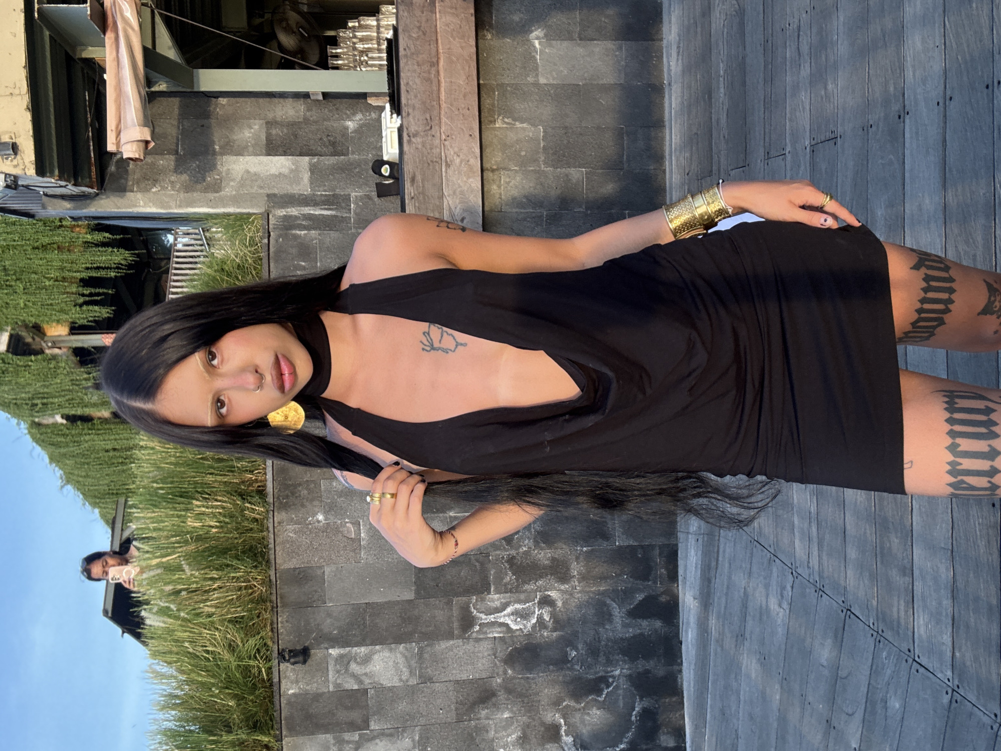 A person with long, dark hair is posing for a photo in an outdoor setting. They are wearing a black, sleeveless dress with a plunging neckline and a dark choker necklace. The person has a gold bracelet on their left wrist and a gold ring on their left hand, and a gold pendant necklace. They also have tattoos on their legs. The background appears to be a patio or deck made of dark wood planks and stone tiles, with some greenery visible. The sun appears to be shining, casting shadows and suggesting it is daytime. The person's gaze is directed towards the camera, with a neutral or slightly curious expression. In the reflection of what appears to be a mirrored surface or window to the left, another person can be seen holding a phone, presumably taking the photo.