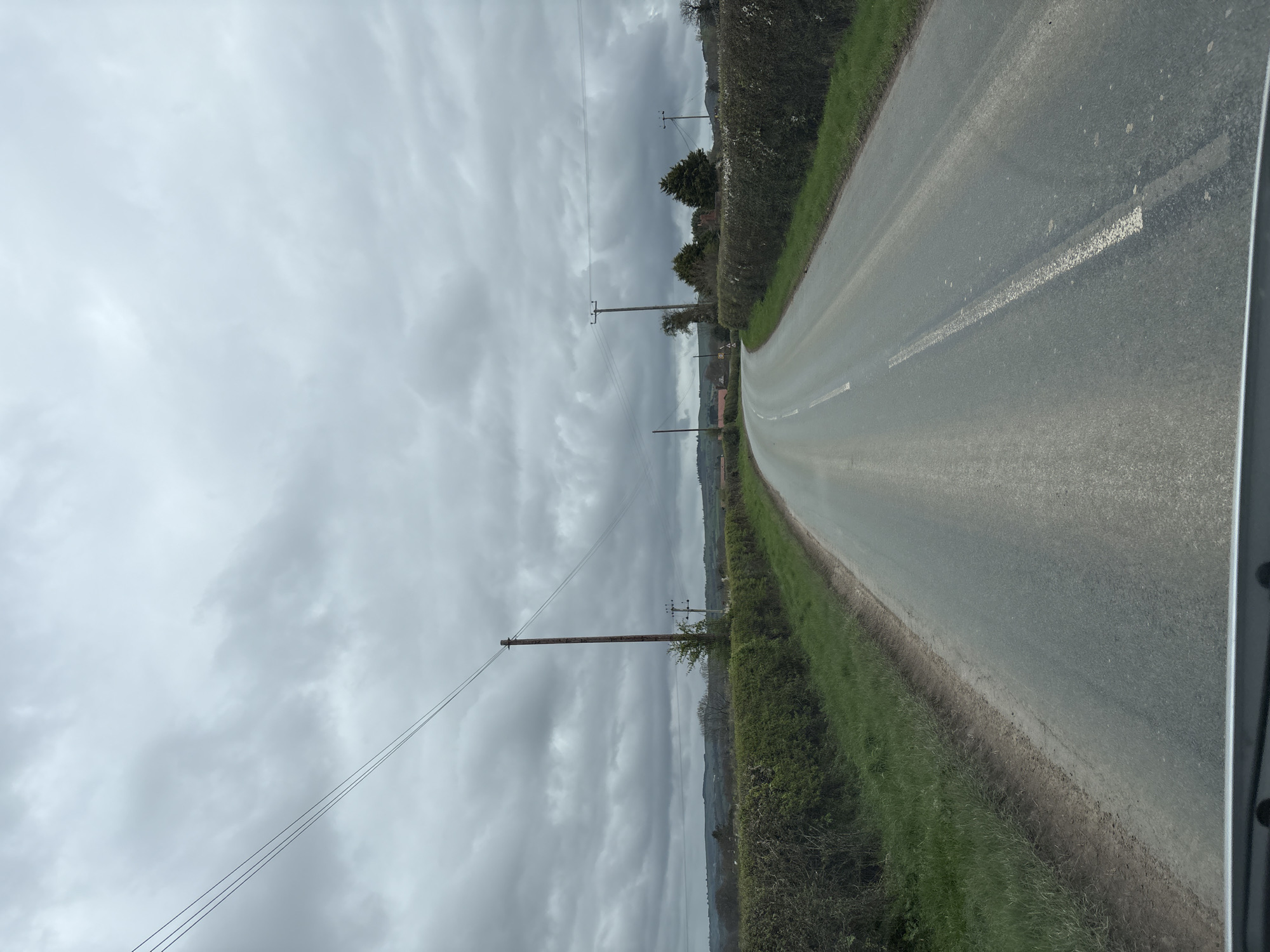 The image depicts a rural road in Longden, United Kingdom, under an overcast sky. The road is asphalt, with a white line marking the center. Hedges and grass line both sides of the road. Utility poles and wires run along the left side of the road. The view suggests a perspective from inside a vehicle.