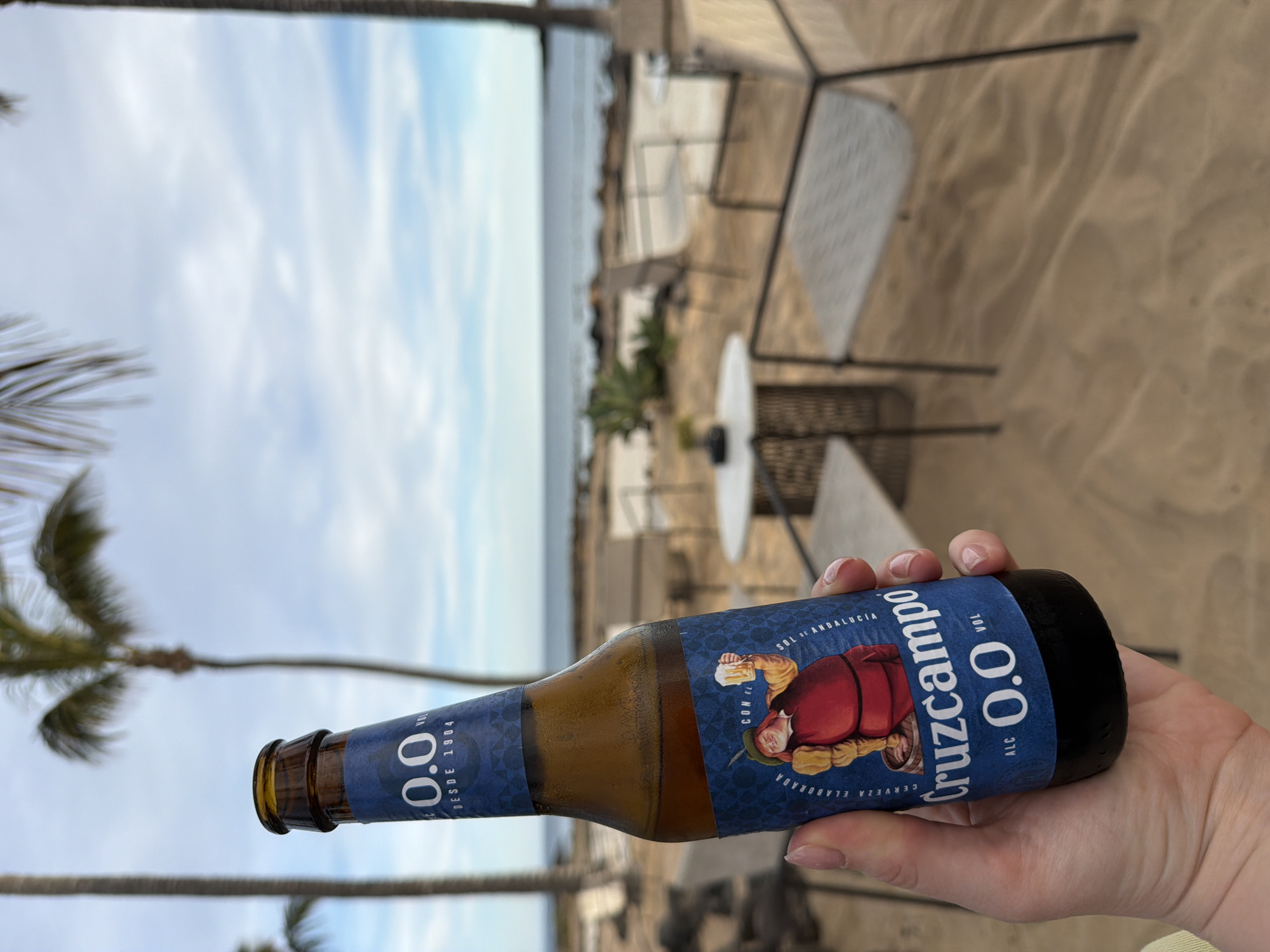 A close-up, rotated image displays a person's hand holding a brown glass bottle of Cruzcampo 0.0 beer. The hand, with neatly manicured nails, firmly grips the bottle, which shows signs of condensation. The bottle's blue label prominently features the brand name "Cruzcampo" and "0.0 ALC VOL," indicating an alcohol-free beverage. An illustration on the label depicts a historical or traditional figure, possibly a monk, holding a beer mug. Additional visible text includes "SOL DE ANDALUCÍA," "DESDE 1904," and "CERVEZA ELABORADA CON EL."
The background reveals an outdoor, coastal setting in Teguise, Spain. The ground beneath consists of soft, light-brown sand. Beyond the hand, several outdoor tables and chairs, constructed from dark metal with light, patterned surfaces, are visible on the sand, suggesting a beachside establishment. In the distance, the horizon separates a calm blue ocean from a bright sky with scattered white clouds. The scene is framed by palm trees, with their fronds visible on both sides of the image. The natural lighting suggests it is daytime, likely during the morning or early afternoon, with clear weather conditions. The overall atmosphere conveys relaxation and leisure in a scenic beach environment.