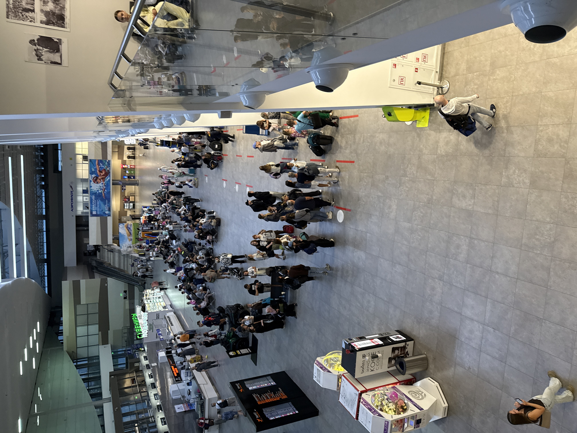 The image shows an aerial view of a busy airport terminal. In the foreground, a woman in light-colored pants and a black top is walking while looking at her phone. To her left are several vending machines, including one with prizes and another labeled "barista ЗЕРНОВОЙ КОФЕ". Further into the terminal, a large crowd of people are seated and standing, many with luggage, suggesting a departure or arrival area. A departure board with flight information is visible on the left. In the background, there are shops, escalators, and more seating areas. The architecture of the terminal is modern with high ceilings and a spacious layout. The overall scene depicts a typical airport environment with travelers waiting. This airport is located in Bolshoye Savino, Russia.