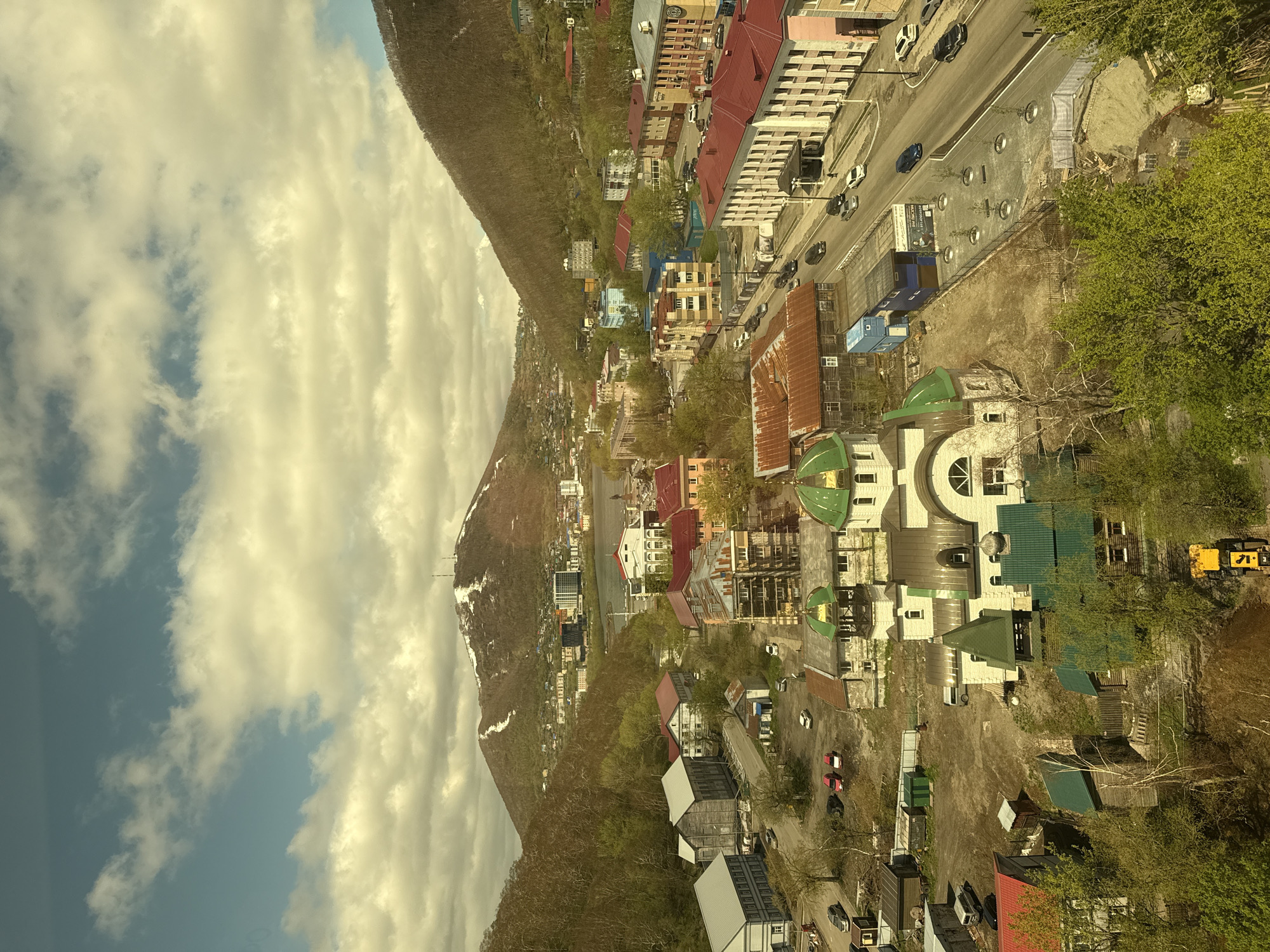 This high-angle, panoramic image captures a vibrant town nestled against the base of a large mountain, under a partly cloudy sky. The perspective suggests it was taken from an elevated position, possibly a cable car or a higher building.
In the background, a prominent mountain dominates the left side of the frame. Its slopes are covered in a mix of trees, some bare and others with fresh green leaves, indicating spring or early summer. Patches of snow are visible near the mountain's summit, suggesting a relatively high altitude or lingering winter conditions.
The midground features a bustling urban landscape. A main road, likely an arterial street, runs through the town with several vehicles, including cars and possibly an SUV, moving or parked. Along the road and extending further into the town, various buildings are visible. These include multi-story residential blocks with distinctive red roofs, smaller houses, and more uniquely designed structures. In the foreground, several buildings stand out with unusual green, dome-shaped roofs and light-colored facades, possibly indicating a specific architectural style or public/religious buildings.
Trees with lush green foliage are interspersed throughout the town, softening the urban environment. In the lower right, an area of undeveloped land or a construction site is visible, with a yellow excavator or similar heavy machinery present, hinting at ongoing development.
The sky is a mix of bright blue and scattered white and grey clouds, suggesting daytime conditions. The overall lighting is bright, casting some shadows and highlighting the details of the buildings and landscape. No discernible human figures or readable text are visible in the image. The scene depicts a quiet moment of daily life in what is likely a town in a mountainous region, consistent with the provided location context of Petropavlovsk-Kamchatsky, Russia.