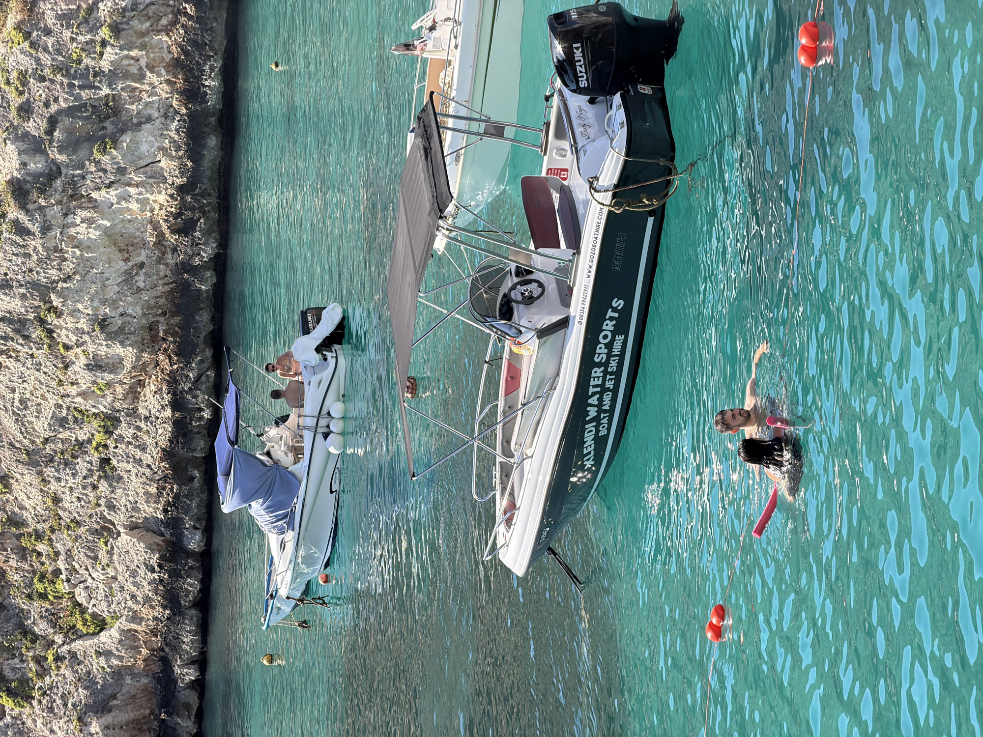 In Xlendi, Malta, a bright, sunny day illuminates a clear, turquoise body of water where two boats are anchored. The larger boat, in the foreground, is a white vessel with a dark green hull inscription that reads "XLENDI WATER SPORTS BOAT AND JET SKI HIRE". It features a black canopy, a steering wheel, and a Suzuki outboard motor. A rope is tied to its stern, connecting to a buoy line with two red buoys visible. In the water near the boat, two people are swimming, one with a pink pool noodle. The second boat, further in the background, is also white with a blue canopy and has three people seated on it. Both boats are positioned near a rocky, sun-drenched cliff face that forms the backdrop of the scene.