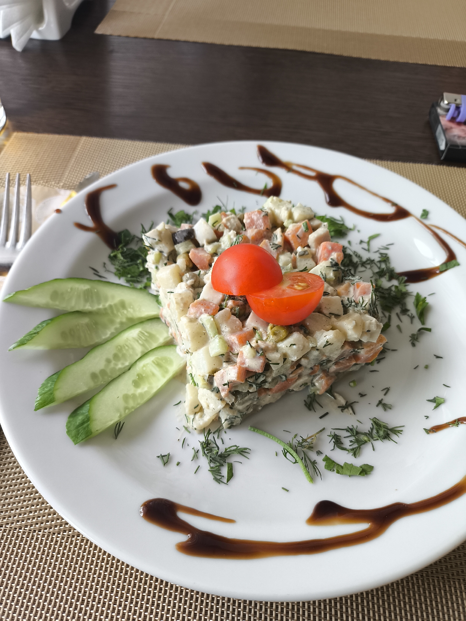 A close-up, eye-level shot features a white plate of salad presented on a brown woven placemat. The salad, a finely chopped mixture with visible pieces of carrot, potato, and possibly meat, is topped with two halves of a cherry tomato. Several slices of cucumber are arranged to the left of the salad, and fresh dill is scattered around the plate. Dark brown sauce is drizzled in a decorative pattern on the plate. In the background, a dark wooden table is visible, along with a white napkin and a cigarette lighter. The scene is set indoors, likely in a restaurant or dining area, and does not provide any specific clues about the city of Taldom, Russia.