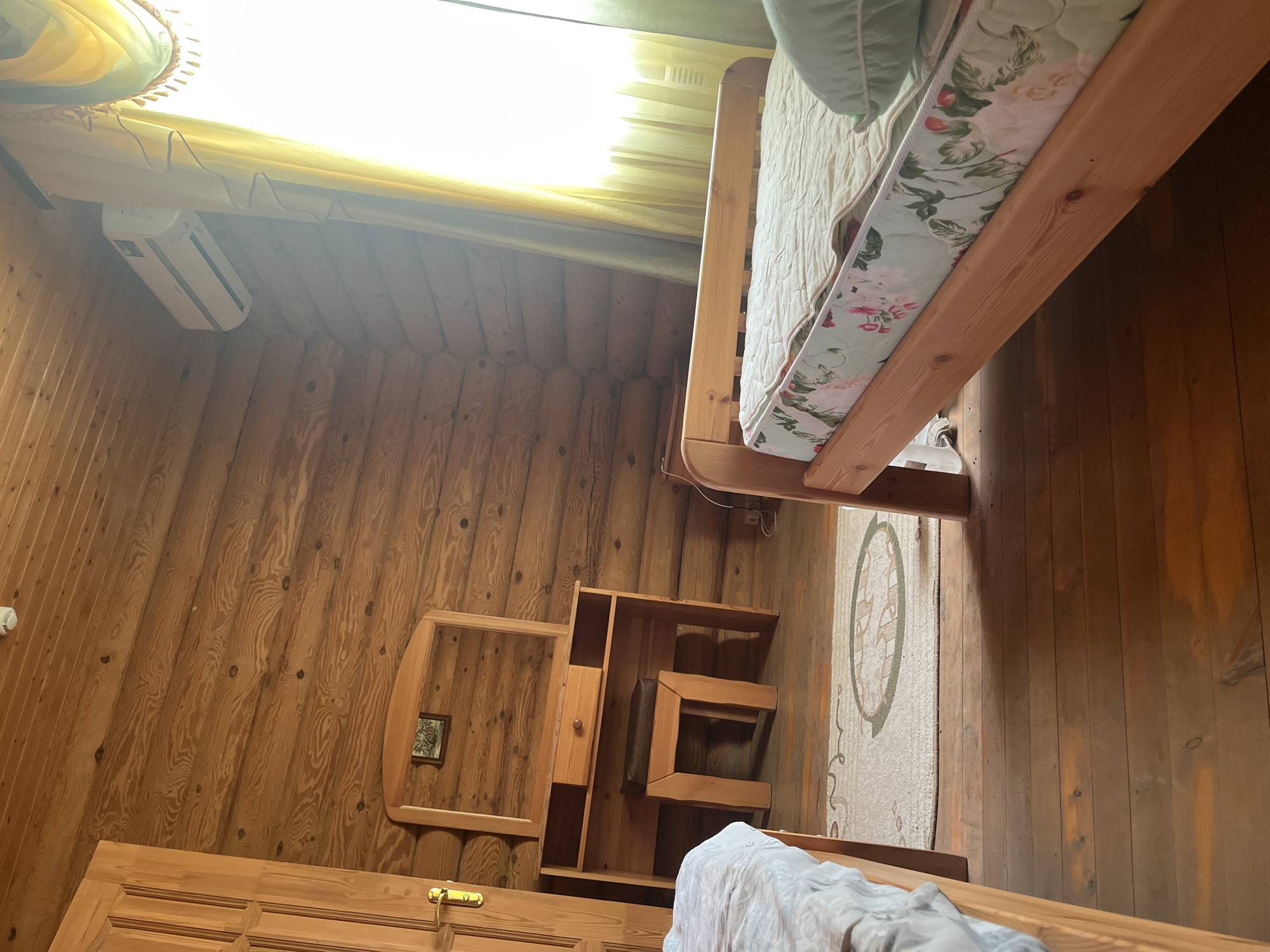The image shows a bedroom in a log cabin. The walls and ceiling are made of light-colored logs. A wooden door with a brass handle is visible on the left. In the center of the room is a wooden vanity with a mirror and a small stool. The vanity has drawers and open shelves. A bed with a floral-patterned mattress is on the right side of the room, and another bed is partially visible on the left. A rug with a circular design is on the floor between the vanity and the bed on the right. An air conditioning unit is mounted on the ceiling on the right side of the room. Yellow and green curtains are drawn across a window on the right. The floor is made of wooden planks. The scene depicts an interior of a room, likely a bedroom, in Koshara, Russia.