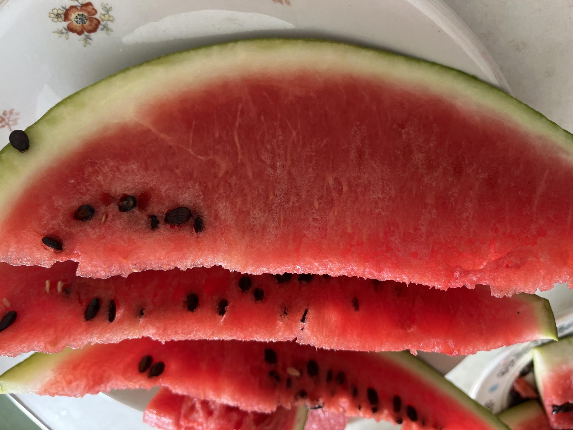 A close-up, high-angle shot showcases several slices of watermelon on a white plate with a floral pattern. The watermelon slices are a vibrant red with black seeds scattered throughout. The rind is a light green. The background is a neutral white. This image was taken in Komsomolskoye, Ukraine.