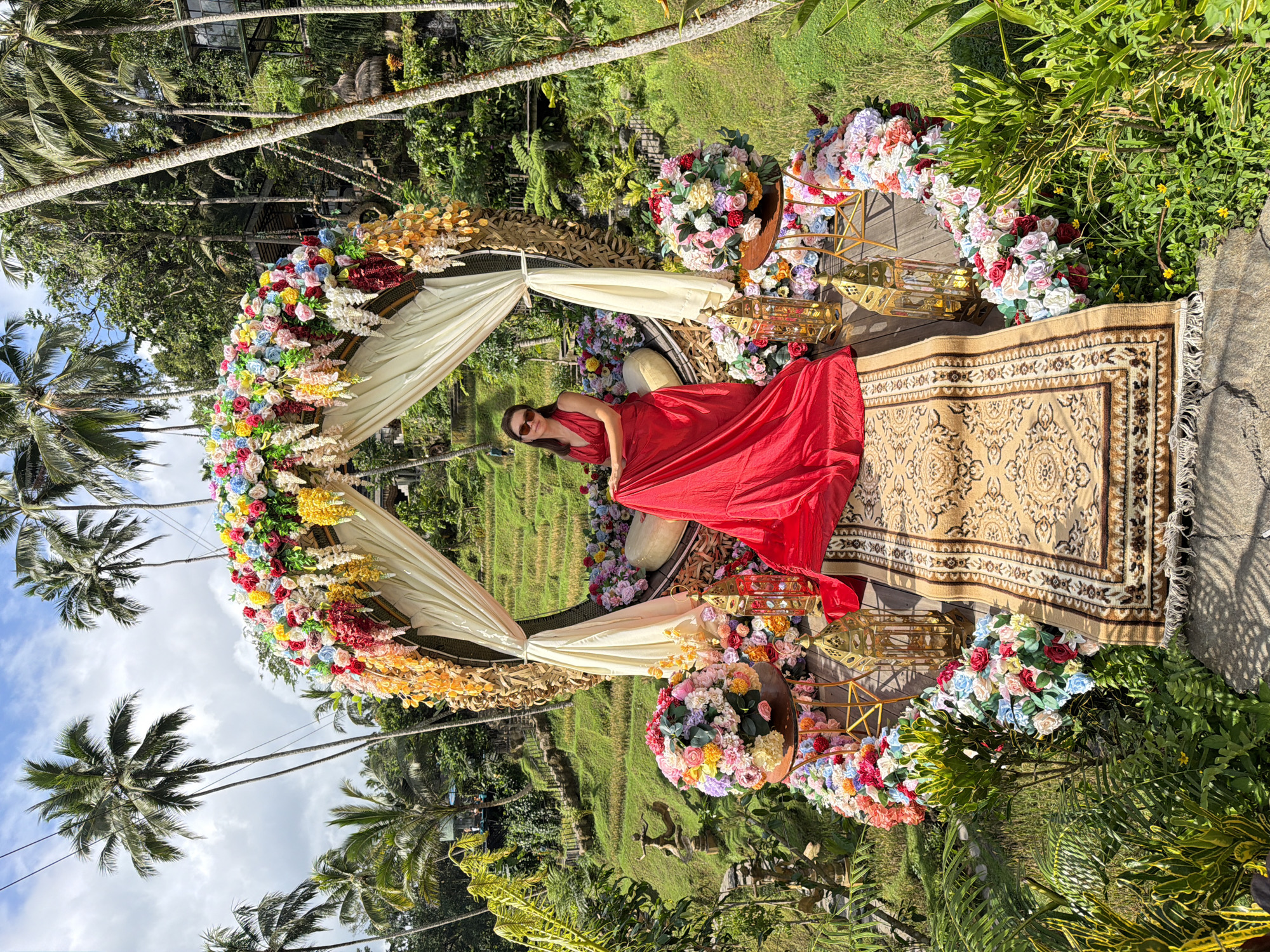 The image shows a woman in a red dress lying on a beige patterned rug within a circular floral archway in Tegal Suci, Indonesia. The archway is decorated with colorful flowers and cream-colored drapery. The rug is on a wooden platform surrounded by more flowers and golden lanterns. Behind the archway are rice terraces and lush greenery. Palm trees are visible in the background against a partly cloudy sky.