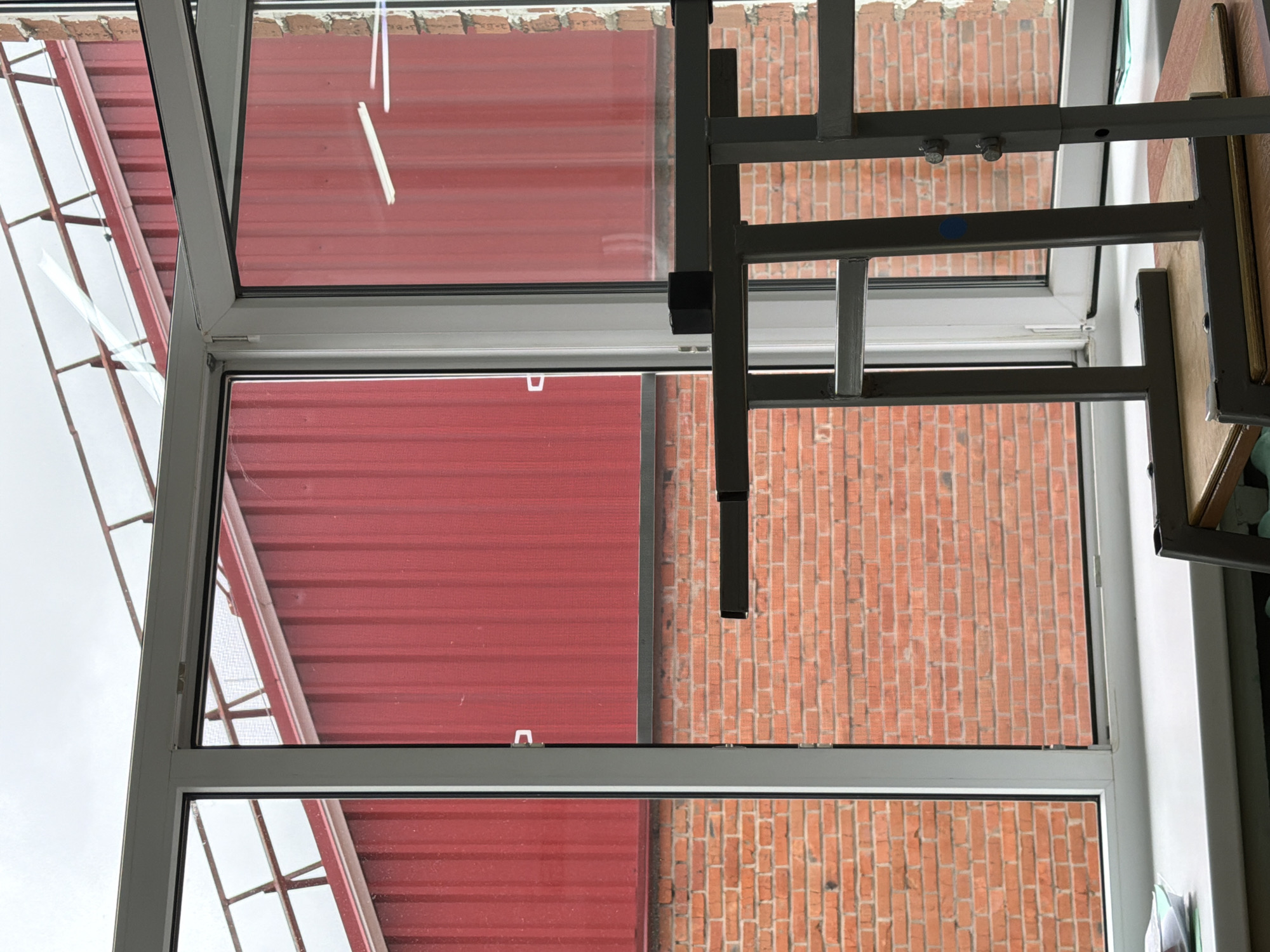 The image shows a view through a window in what appears to be an interior space. On one side of the window is a red, corrugated metal structure, possibly a roof. On the other side is a brick wall of an adjacent building. A gray metal frame, likely part of furniture or equipment within the interior space, is partially visible within the window frame. The sky is overcast. The location of Noviy, Russia is not ascertainable from the image.