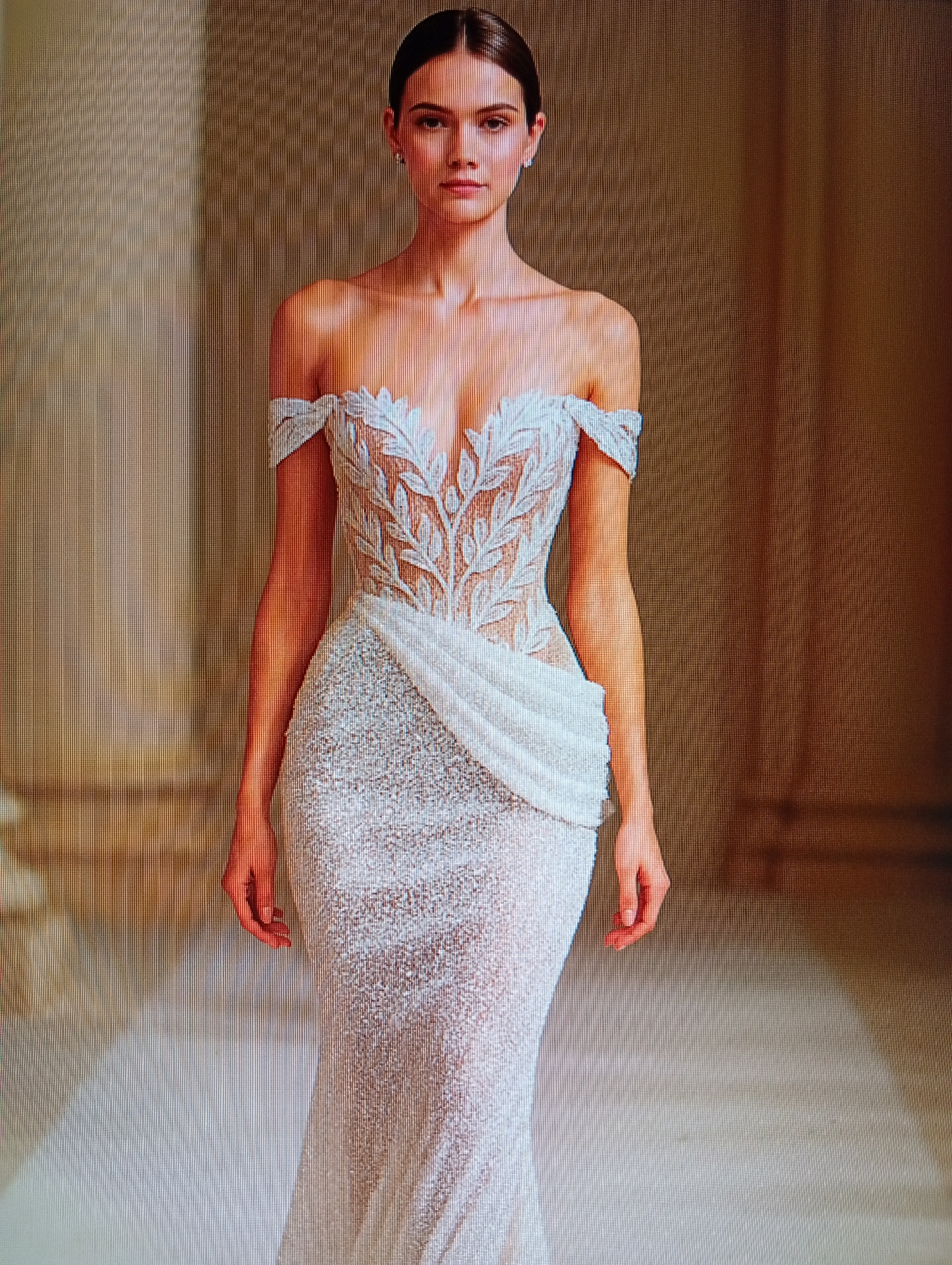 A full-body, eye-level, slightly low-angle, medium shot captures a fashion model posing in a white, off-the-shoulder, floor-length gown with intricate beadwork. The gown features a plunging neckline with leaf-like embellishments extending over the bust and upper bodice. A draped element is present on the right hip, creating a cascading effect. The skirt of the dress is also heavily beaded, tapering towards the hem. The model has her hair pulled back and is wearing subtle earrings. She stands in what appears to be an indoor venue with architectural columns in the background, illuminated by soft lighting that casts a warm glow. The environment suggests a runway or photoshoot setting.