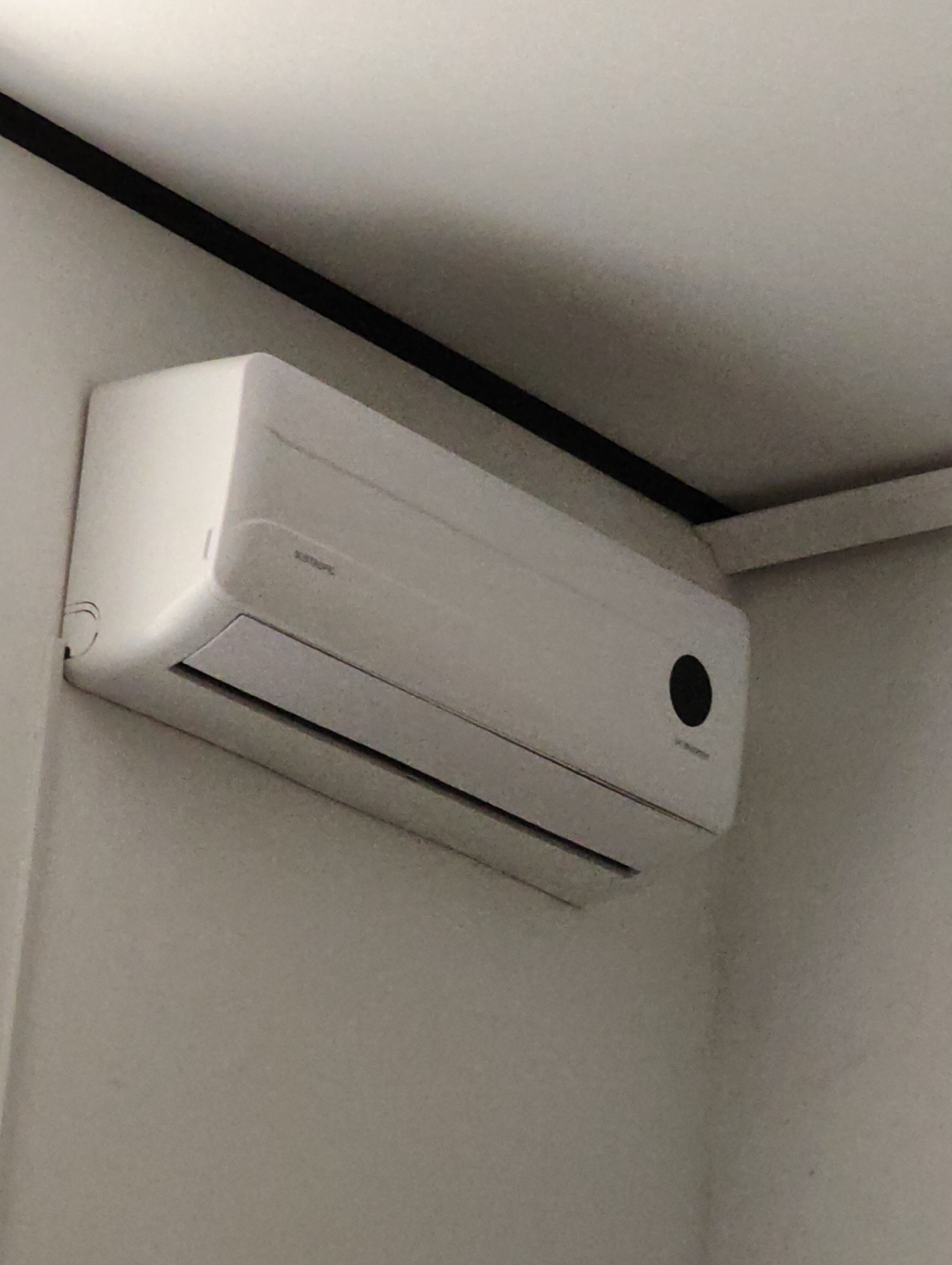 A white indoor air conditioning unit is mounted high on a light-colored wall, near the ceiling. A black line runs along the seam where the wall meets the ceiling. The air conditioner has a sleek, modern design with a prominent black circular indicator on its right side. A louvered vent is visible at the bottom of the unit. The image is taken from a low-angle perspective, looking up at the air conditioner. The scene suggests a residential or office interior. No people are present in the image. The overall impression is one of a clean, minimalist environment. The location in the city of Komsomolsk-on-Amur, Russia is not discernible from the image alone.