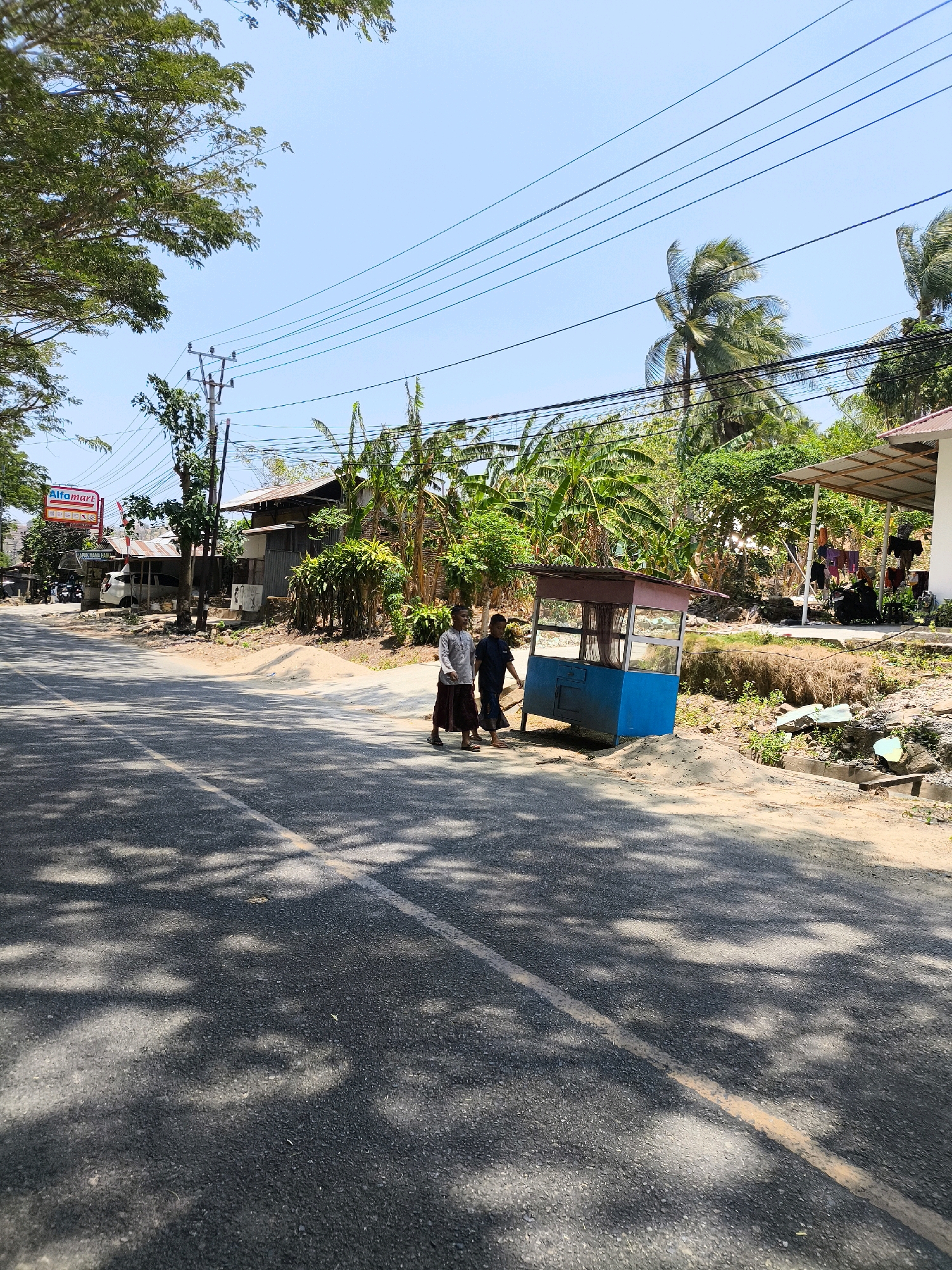 A street-level view captures a sun-drenched, tropical scene in an Indonesian town. The asphalt road, marked with a yellow line, is partially shaded by the dappled light filtering through overhead trees. Two young individuals, a boy and a girl, walk along the side of the road, their casual attire suggesting a warm climate. A small, blue and red kiosk stands nearby, a common sight in many Asian towns, suggesting a commercial area. In the background, simple houses with corrugated tin roofs and lush vegetation, including banana trees and palm trees, are visible. Power lines crisscross the sky, a common element in developing regions. The overall impression is one of a peaceful, everyday scene in a rural or semi-urban Indonesian setting.