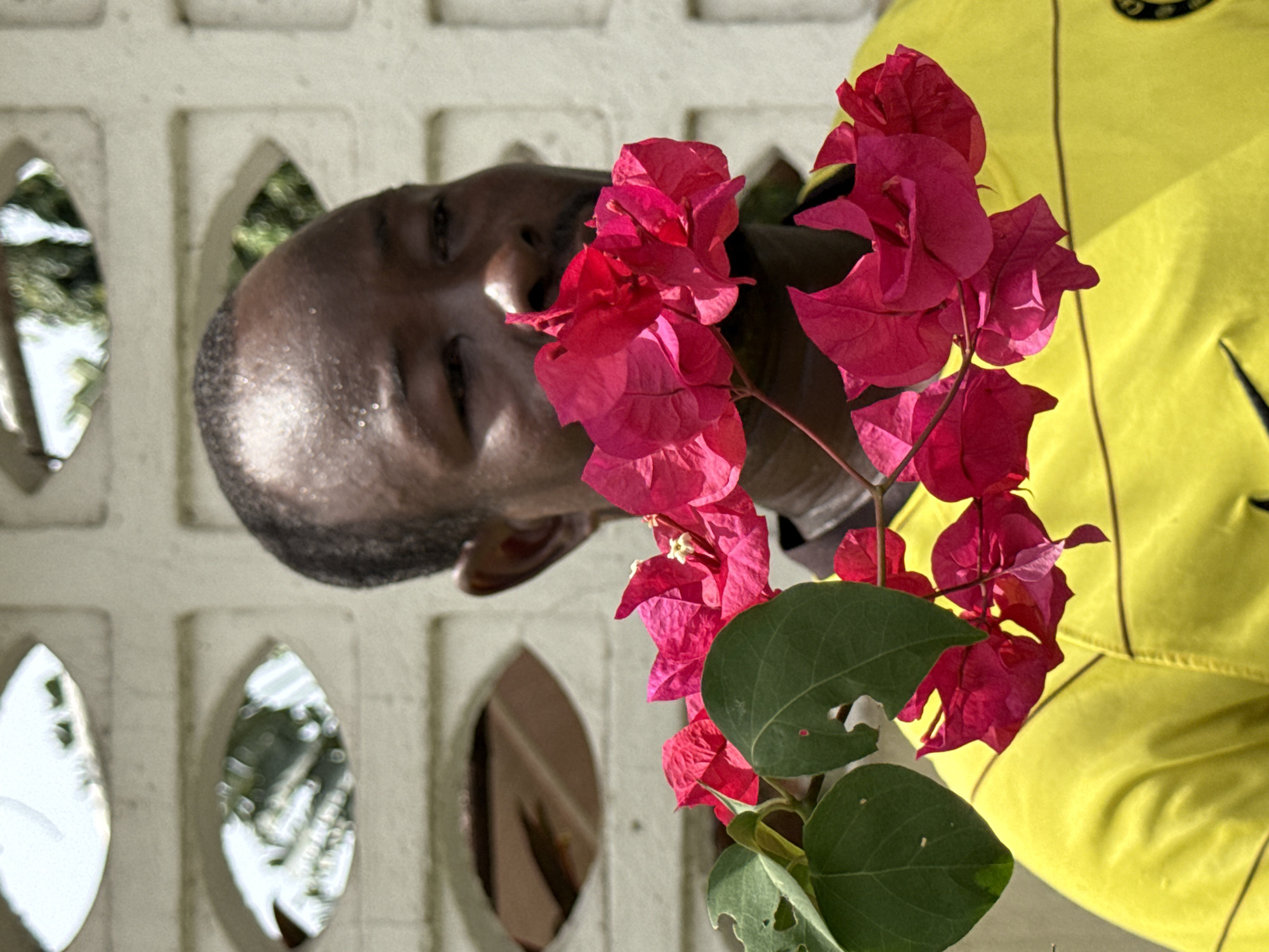 An outdoor portrait captured in Lagos, Nigeria, features a dark-skinned man with short hair, positioned on the left side of the frame. He is looking slightly upwards and to the right with a serene or gentle expression, his face illuminated by bright daylight. Directly in front of and to the right of his face is a vibrant cluster of magenta bougainvillea flowers with green leaves, partially obscuring his lower face and left shoulder. The man is wearing a bright yellow garment, possibly a shirt or jacket, which is visible on the right side of the image. This garment has subtle dark stitching lines and a dark, circular logo on the upper chest, though the text within the logo is not clearly legible.
The background consists of a light-colored wall with distinctive decorative cutouts, which appear to be geometric, possibly leaf-shaped or oval, and are characteristic of concrete screen blocks. Through these openings, blurred green foliage and bright, sunny sky are visible, indicating an outdoor setting. The strong, direct sunlight creates prominent highlights and shadows, confirming it is daytime. Some of the bougainvillea leaves show minor imperfections, such as small holes. The overall atmosphere of the image is bright and peaceful.