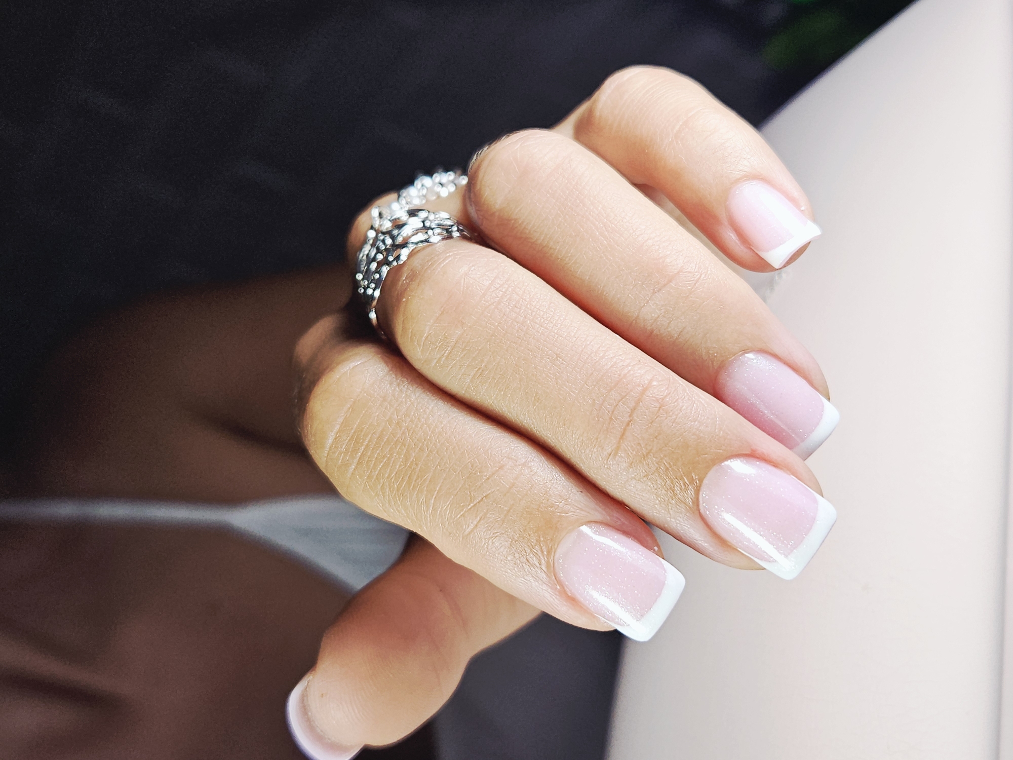 A close-up shot shows a person's hand with a silver ring on the ring finger. The fingernails are neatly manicured with a French tip design, featuring a light pink base and a white edge. The hand is posed against a background that transitions from dark to light, with hints of white clothing visible.