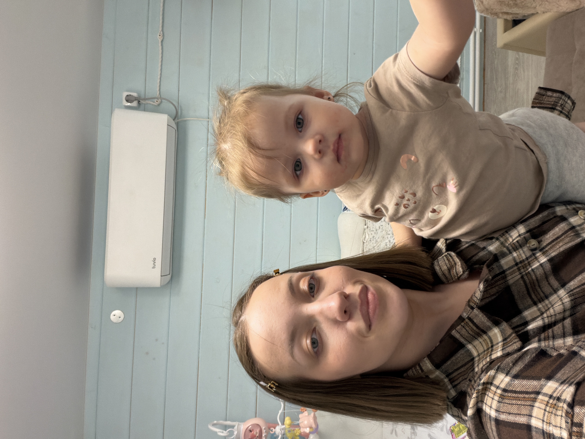 The image depicts a fair-skinned woman with brown hair in a plaid shirt, posing for a selfie with a fair-skinned toddler with blonde hair in a brown shirt. The woman is looking directly at the camera, while the toddler is looking slightly off to the side. The background shows a light blue wall with vertical panels, an air conditioning unit, and a baby mobile hanging from the ceiling. The location cannot be determined from the image.