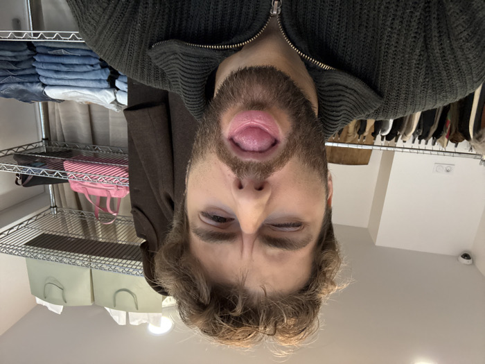A bearded male, appearing in his 20s or 30s with brown hair, winks his right eye and extends his tongue towards the camera, filling the majority of the frame. He wears a dark green, textured, collared sweater with a zipper. The scene is an indoor retail or storage space located within Paris, France. In the background to the left, a white wall displays two electrical outlets and a white dome-style security camera mounted in the upper corner. Below, a metal clothing rack holds various dark and patterned garments. To the right of the subject, a metal wire shelving unit is visible. The top shelf contains two light green shopping bags. The middle shelf holds a bright pink handbag with white diagonal stripes, next to a darker bag. The bottom shelf is stacked with neatly folded denim clothing in various shades of blue.