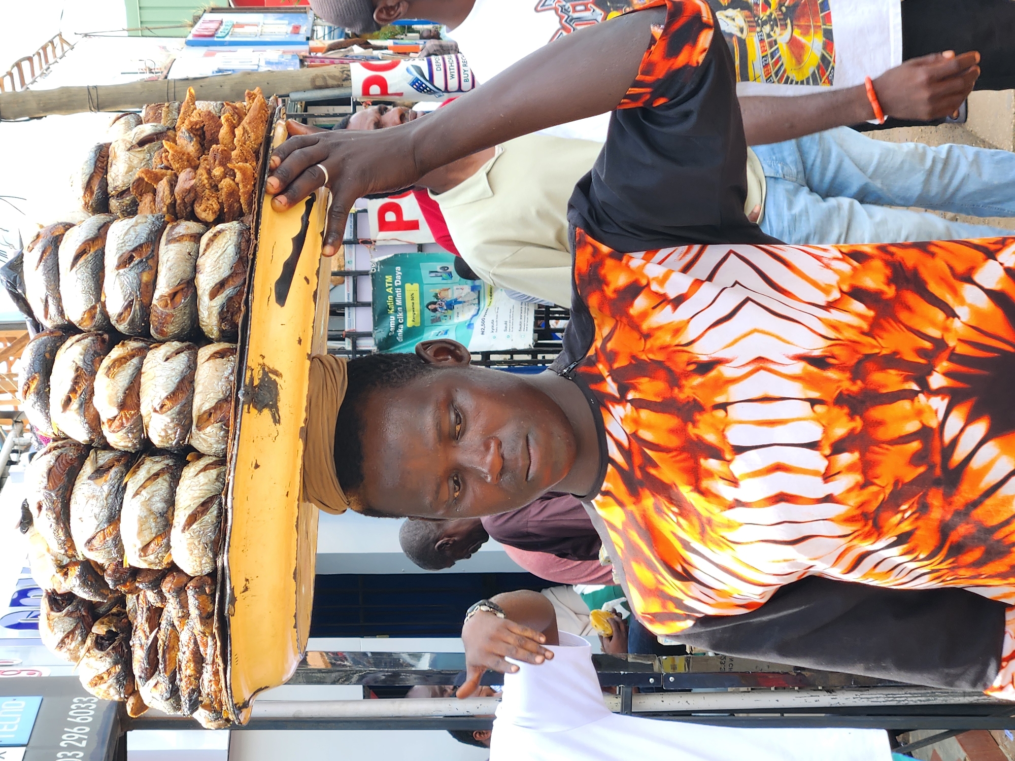 A young Black man, a vendor, balances a large tray of fried fish and chicken on his head. He wears a black and orange tie-dye shirt. His left hand, adorned with a ring and a red bracelet, grips the tray from the side, while his right hand is not visible. He is looking down, seemingly focused on his balance or on potential customers. Behind him, other people are visible, including a man in a tan shirt and blue jeans, and another wearing a white t-shirt with a colorful graphic. The scene appears to be a street market in a city, with buildings and signs in the background, suggesting an urban environment. The lighting is bright, indicating it is daytime.