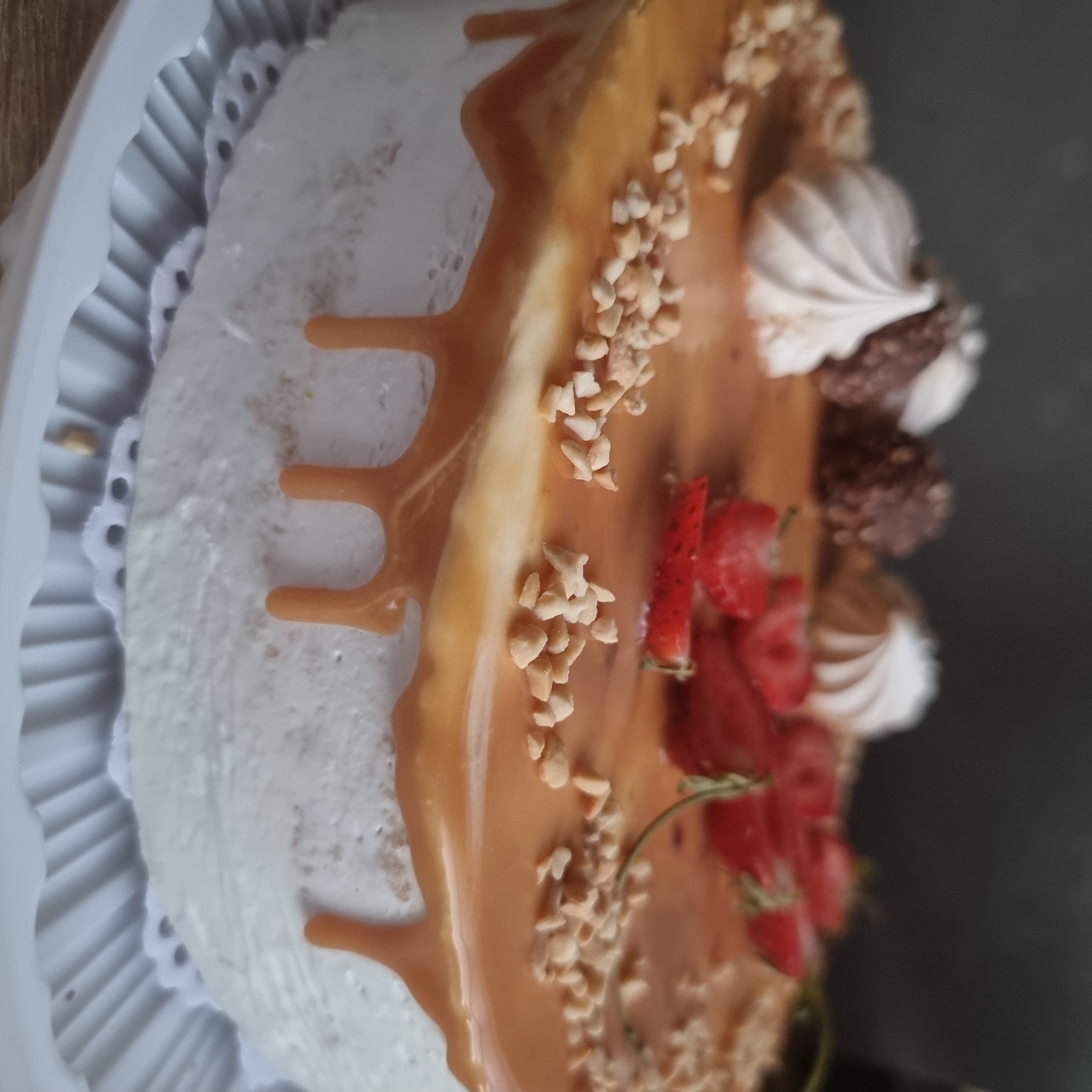 The image shows a close-up, slightly blurry, overhead view of a caramel-drizzled cake. The cake is frosted with white icing and topped with scattered chopped nuts, several sliced strawberries, and three meringue kisses. In the background, the environment is a dark, blurred wall, and the cake rests on a white decorative serving platter. There is no indication of the city of Aktobe, Kazakhstan, in this image.