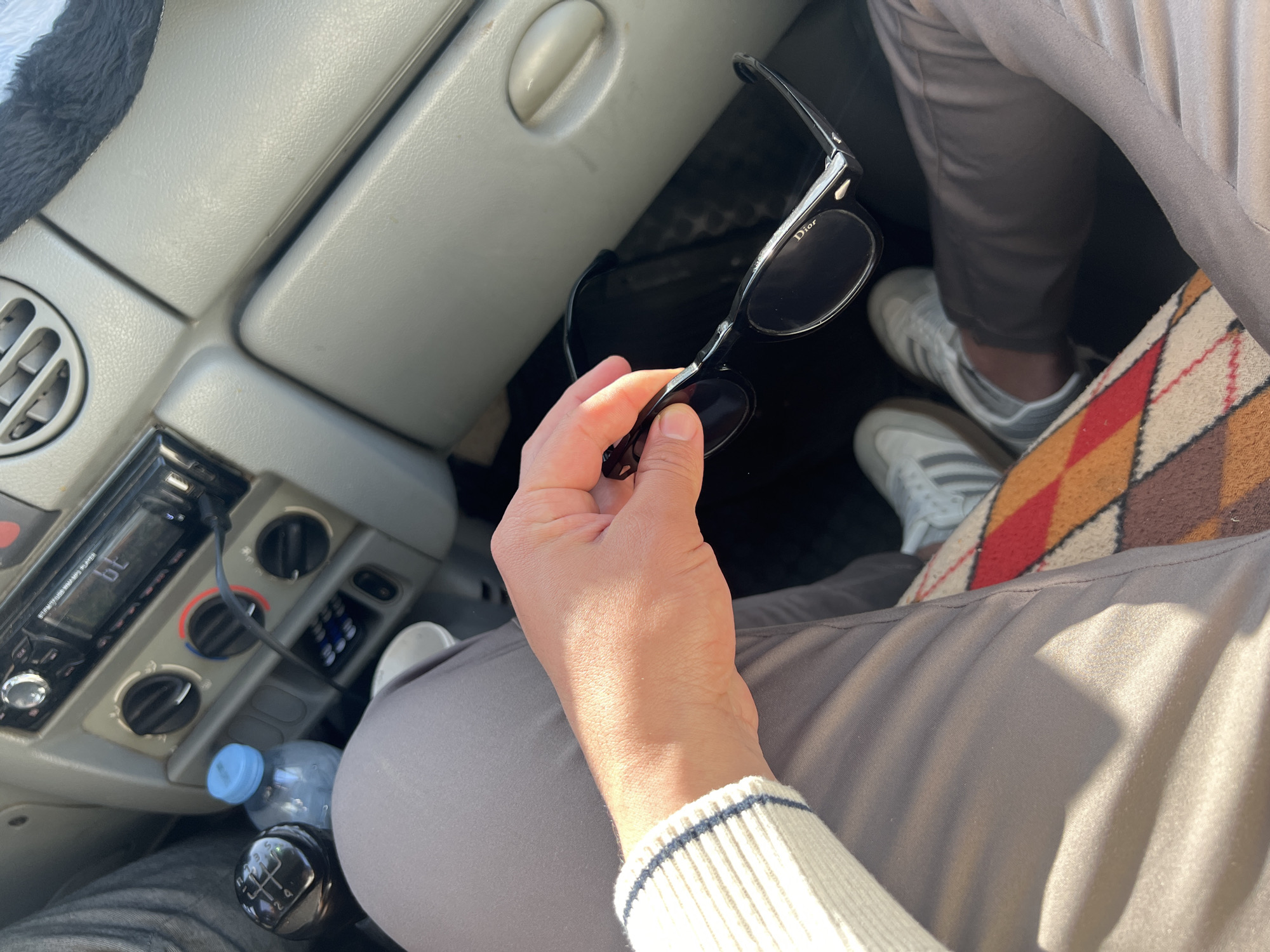 Inside a vehicle, an individual holds a pair of black sunglasses in their hand. The car's interior is visible, including the dashboard, radio, climate controls, gear stick, and a water bottle. Parts of the individual are captured, including a light-colored sweater, grey trousers, and white sneakers. The location is indicated as Martil ⵎⴰⵔⵜⵉⵍ مرتيل, Morocco, based on given information.