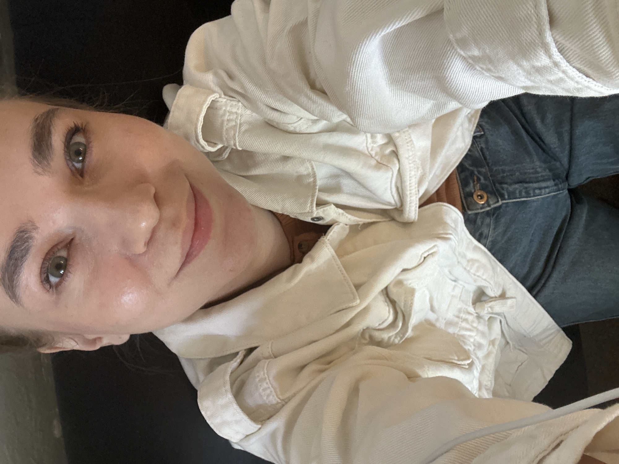 A close-up, angled selfie of a fair-skinned woman with light brown hair. Her face is oriented to the left of the frame, with her nose centrally located. She has light eyes and full lips. She wears a light-colored denim jacket. To her right are blue denim jeans with a visible button. A dark surface, likely seating, is behind her head. The location in the city of Chaikovsky, Russia, is not apparent from the image.