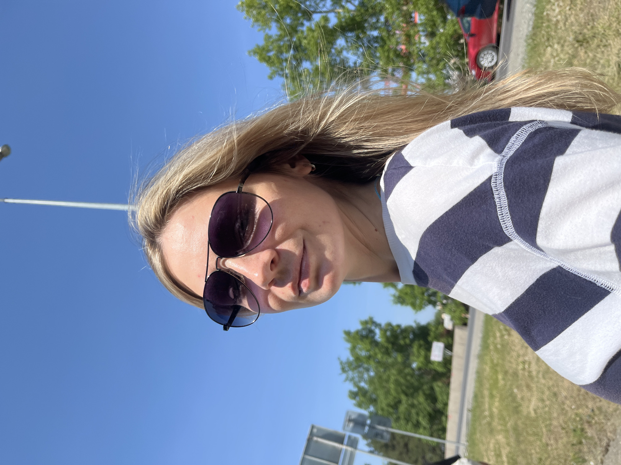 A medium shot of a woman wearing sunglasses and a horizontally striped white and dark blue shirt. The background includes a clear blue sky, greenery, and a glimpse of a red car. A street light is visible in the distance. The location may be Chaikovsky, Russia.