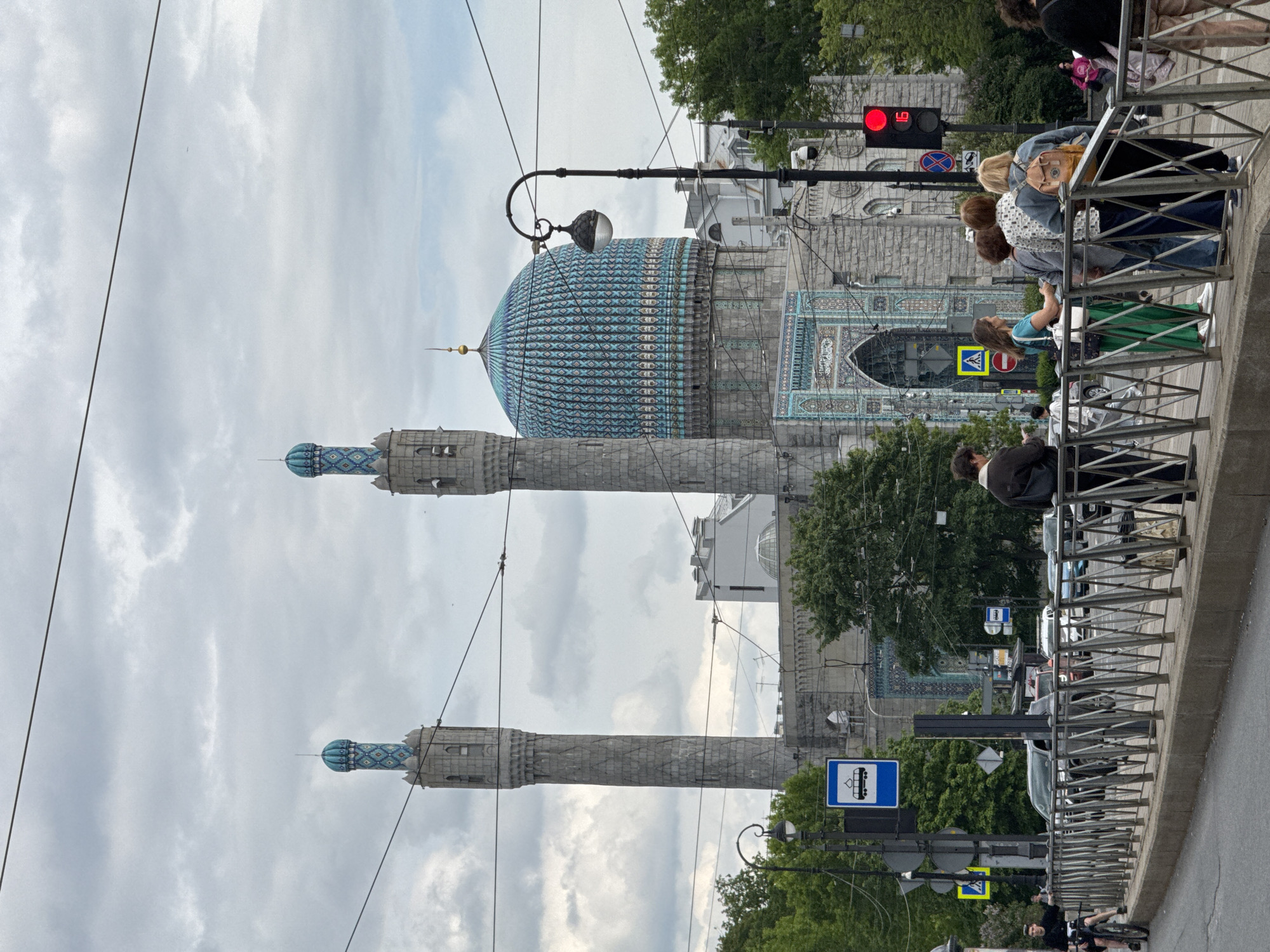 This image captures a daytime urban street scene in Saint Petersburg, Russia, centered around the distinctive Saint Petersburg Mosque under an overcast sky. The mosque, a prominent architectural subject, features a large, intricately tiled blue dome with a golden finial, flanked by two tall, slender minarets topped with smaller blue domes. Its facade is adorned with ornate turquoise and white tilework.
In the foreground, a metal pedestrian railing runs horizontally, separating the viewer's vantage point from a busy sidewalk and street. Several people are visible: some stand near the railing, appearing to observe or wait, while others move along the sidewalk. One individual is seen cycling further down the street. The people are casually dressed, and their expressions are not discernible.
The street is lined with parked and moving cars. Overhead, a street lamp extends into the frame, and multiple power lines crisscross the cloudy sky. A red traffic light, prominently displaying the number "0" (likely indicating time remaining), hangs above the street. Various road signs are visible, including a blue square sign with a white bus/trolleybus icon, a yellow triangular pedestrian crossing sign, and a red circular "no entry" sign. Green trees are interspersed throughout the scene, adding natural elements to the urban landscape. The soft, diffused lighting suggests a cloudy day, possibly late morning or afternoon.