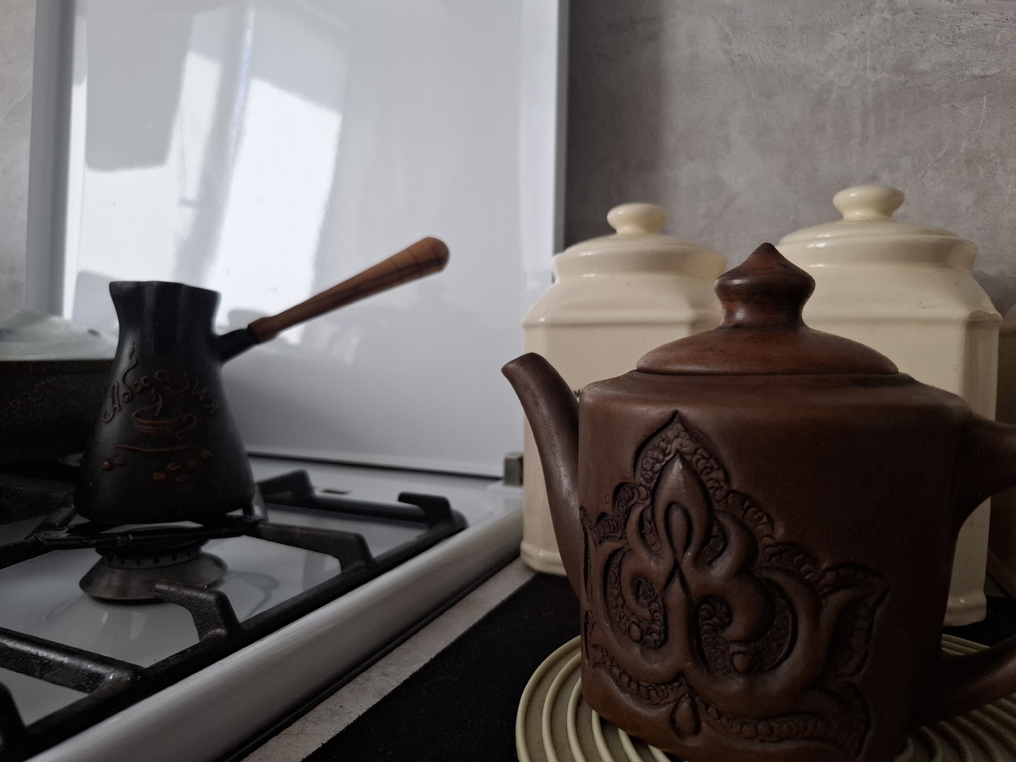 The image shows a kitchen scene. On a white gas stovetop, a black Turkish coffee pot with a wooden handle sits on one of the burners. To the right, a brown ceramic teapot with intricate raised designs rests on a circular trivet. In the background, two lidded jars stand near a gray wall. The location is Амзя, Russia.
