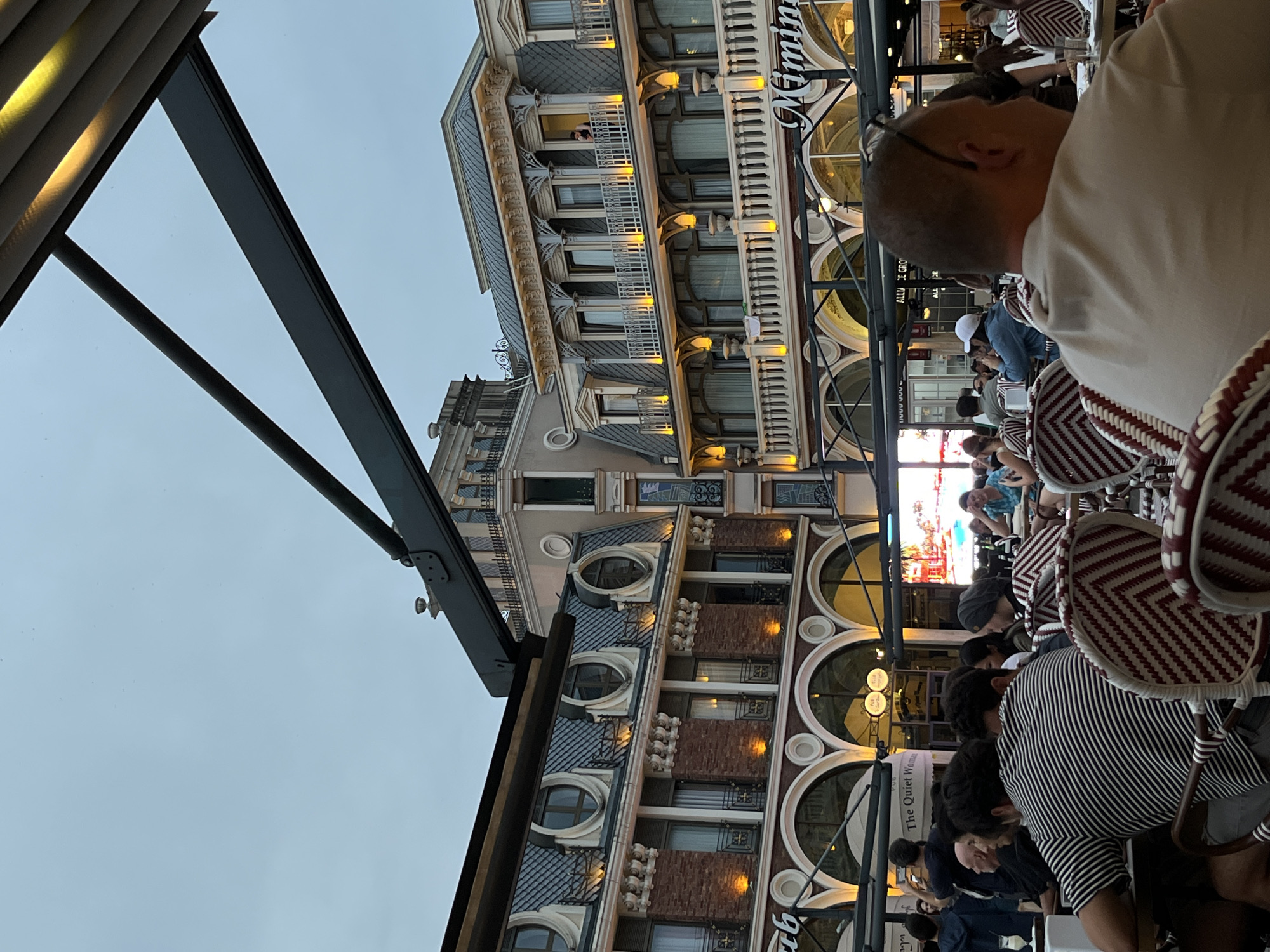 The image depicts an outdoor seating area of a restaurant with people dining. In the background, there are ornate buildings, characteristic of the architecture found in Batumi, Georgia. The buildings feature multiple stories with balconies, decorative cornices, and large arched windows. There are signs visible, one of which reads "The Quiet Woman." People are seated at tables, some are interacting, and others appear to be looking at their phones or the surroundings. The lighting suggests it is either dusk or nighttime, with artificial lights illuminating the buildings and the seating area. The overall atmosphere seems relaxed and social, typical of a restaurant environment in a city.