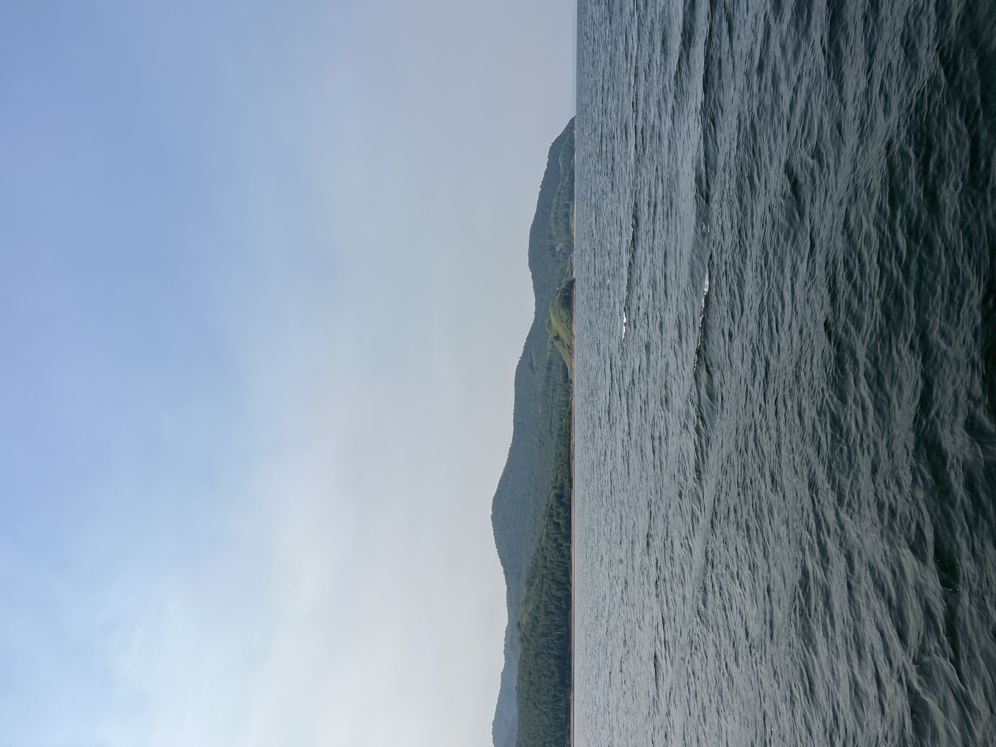 The image shows a wide body of dark water with moderate ripples, stretching towards a distant shoreline. The shoreline is characterized by rolling green hills covered in dense forest. The sky above is a pale blue with hazy clouds. The overall scene depicts a natural landscape, likely a lake or a wide river, given the calm, expansive water and the forested hills. The location is described as being in the city of Novoselovo, Russia, suggesting it is a view from the Krasnoyarskoye Reservoir or a similar body of water within the region. There are no people or man-made structures immediately visible in the frame.