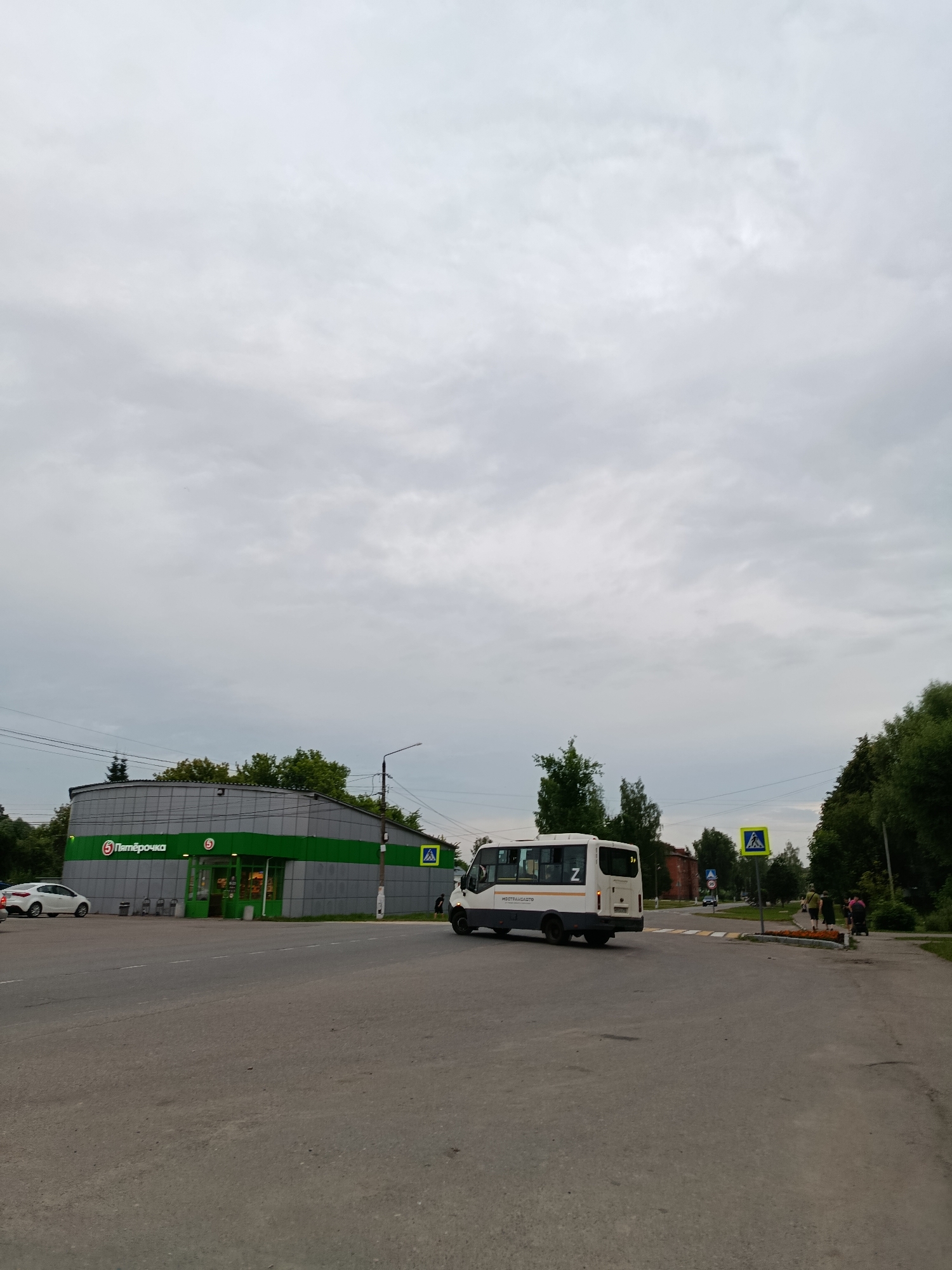 This media file depicts a street scene in Vereya, Russia, under an overcast sky. The time of day appears to be late afternoon or early evening, given the diffused lighting and cloud cover.
The main subjects include a white bus with the letter "Z" on its rear, a white car parked near a building, and several pedestrians crossing a street. The bus has text on its side, though it is partially obscured.
The setting is a commercial area featuring a "Пятёрочка" (Pyaterochka) grocery store. The store has a distinctive green and grey facade with large windows and an entrance. Adjacent to the store are streetlights and trees. A pedestrian crossing is visible, marked by a yellow diamond-shaped sign with a pedestrian symbol. Further down the street, other vehicles and buildings can be seen, along with more trees lining the road.
The overall mood of the image is mundane, capturing an ordinary day in a suburban or small-town environment. The activity taking place is typical urban life: people are commuting and shopping. The weather is clearly cloudy, suggesting a potentially cool or damp atmosphere. The presence of the "Z" symbol on the bus might indicate a political or social context, but without further information, it's purely an observation of a visible detail.