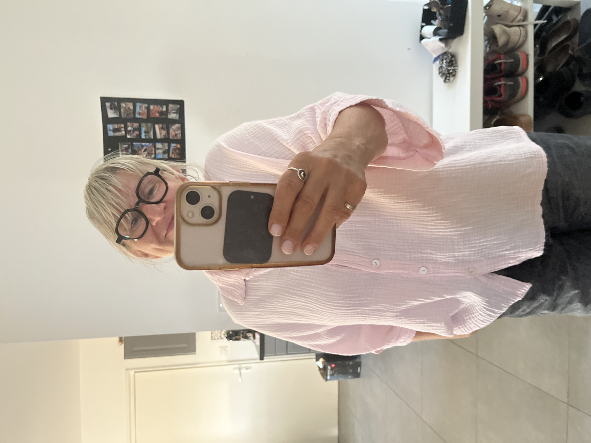 A woman stands in what appears to be a hallway or entryway, taking a selfie in a mirror. She has blonde hair and is wearing large, black-rimmed glasses. She is dressed in a light pink, crinkled fabric shirt and dark grey pants. On her left hand, she wears two silver rings. Her phone, held in her right hand, is a light-colored case with a black object attached to the back. In the background, a white door is visible on the left, with a grey cabinet mounted on the wall above it. To the right of the woman, a framed collage of small photographs is hanging on the wall. Further to the right, a white shelf unit holds various items, including a black container with toiletries and a pair of shoes. The floor is tiled with large, square, light-colored tiles. The setting is likely a residential interior.