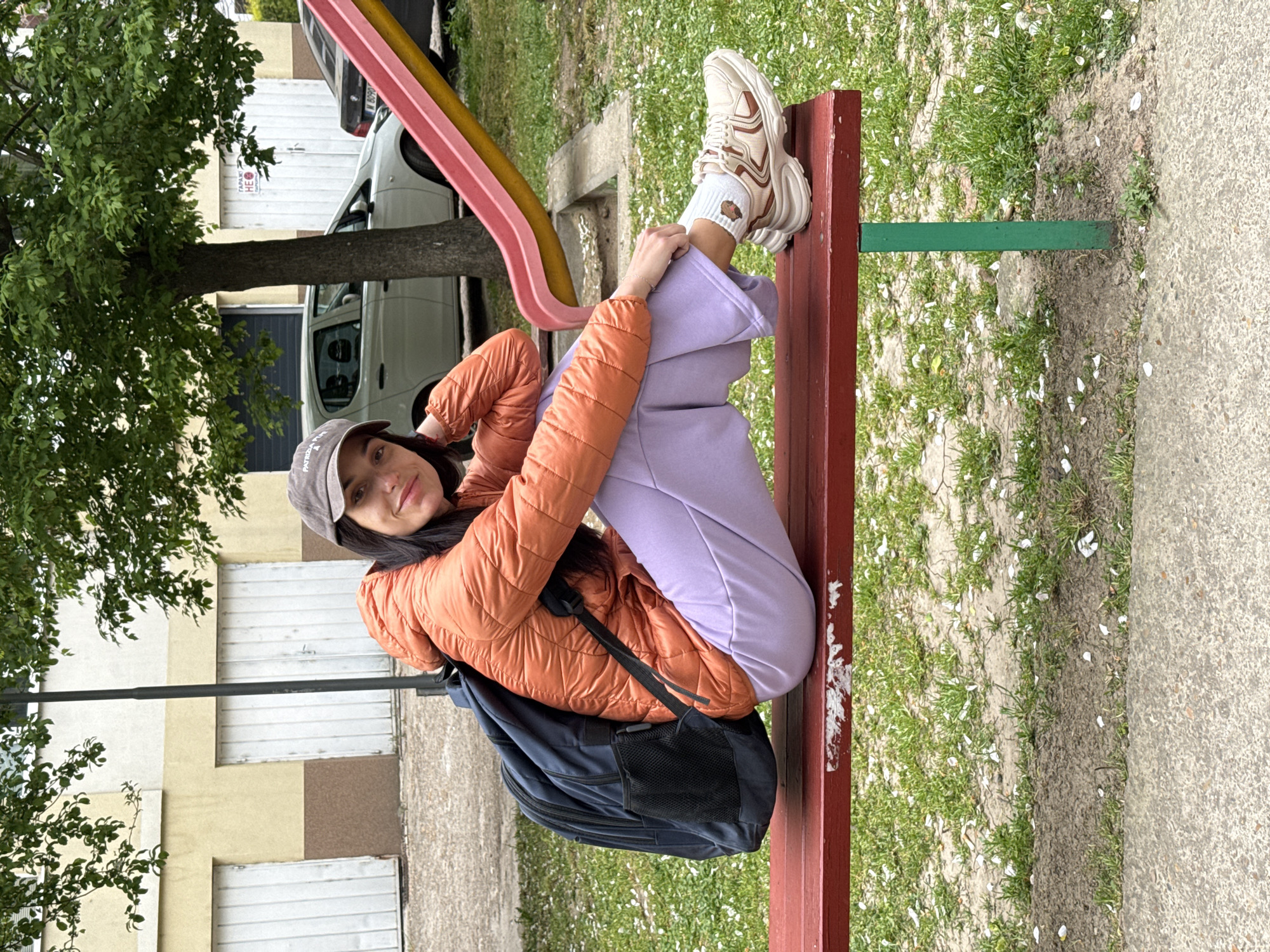 A young woman is seated on a red park bench outdoors. She is wearing a grey baseball cap, an orange puffer jacket, lavender sweatpants, white socks, and beige sneakers. She has a navy blue backpack on the bench beneath her. A colorful playground slide is visible in the background, along with a parked car and an apartment building with closed white shutters. The ground is covered in grass and fallen white flower petals. The image was taken in Pleven, Bulgaria.