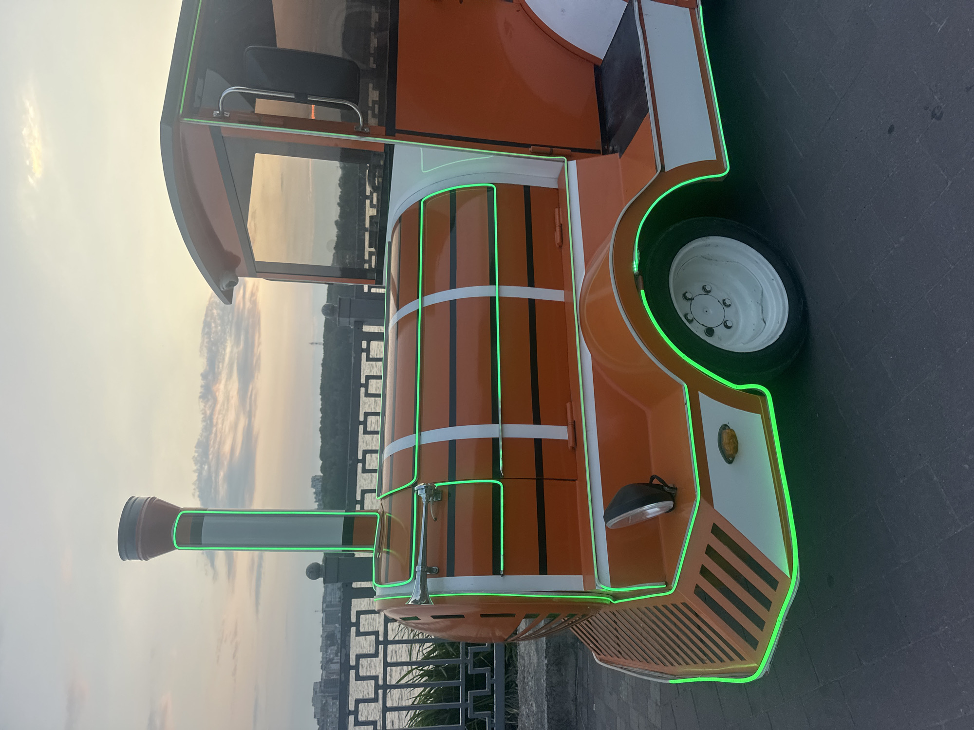 The image shows a bright orange novelty train, likely a tourist attraction, parked on a paved walkway next to a body of water. The train is adorned with vibrant green neon lights that outline its distinctive features, including the windows, doors, and the front "smokestack." The train's design is playful, with a rounded front and a visible driver's cabin with a dark seat.
In the background, a body of water, possibly a river or lake, is visible, with trees lining the opposite bank. A decorative metal railing separates the walkway from the water. The sky is overcast with soft, diffused light, suggesting either early morning or late afternoon. The clouds are tinged with the warm hues of a sunset or sunrise, creating a gentle atmosphere. The overall setting appears to be a park or promenade area, a common place for such tourist vehicles. No people are visible in the frame, and no text is present. The image was taken in Ternopil, Ukraine.