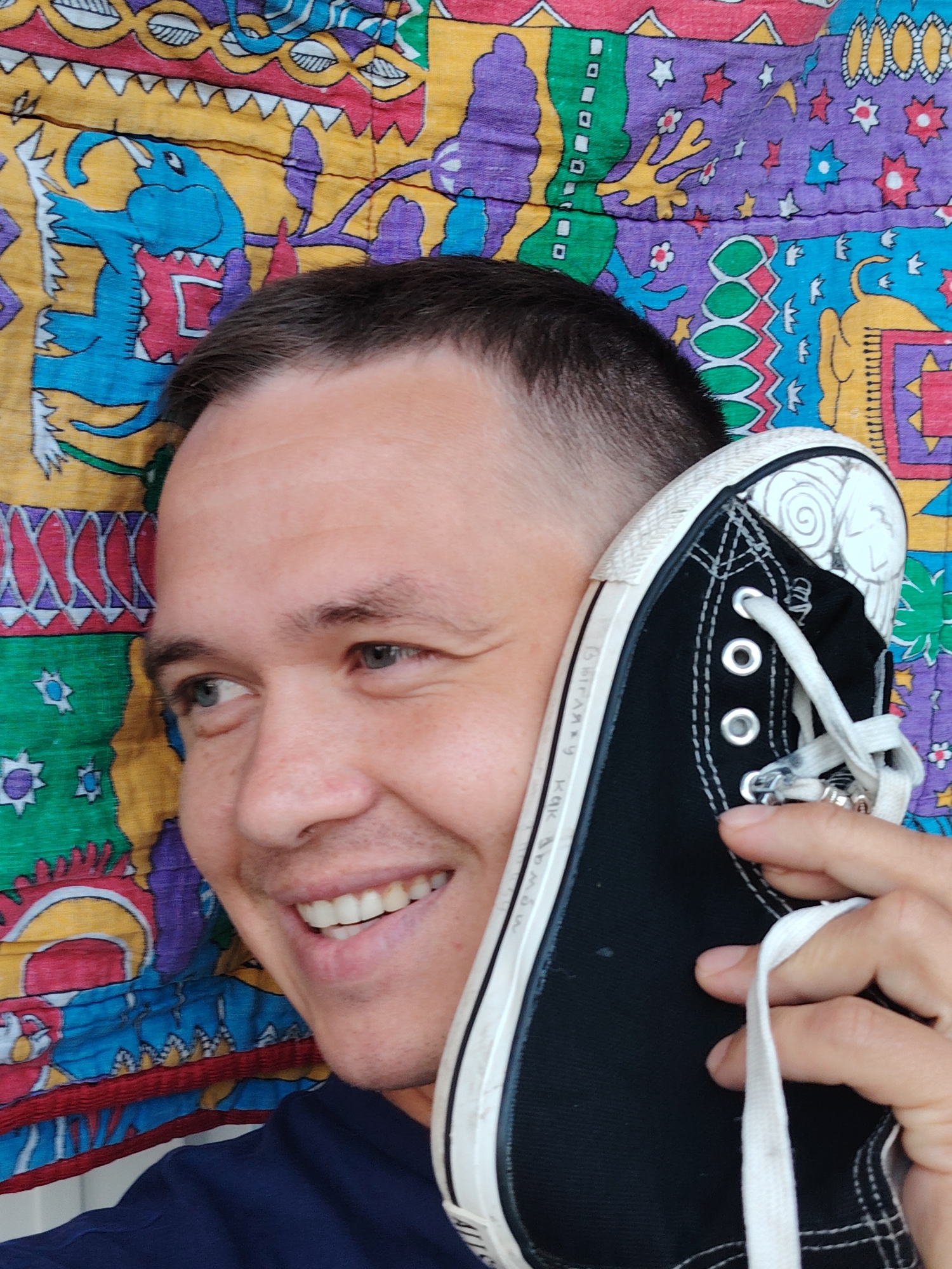 A close-up, eye-level shot shows a man holding a black Converse sneaker to his right ear, as if making a phone call. He is smiling and looking to his left. The man has short brown hair, blue eyes, and light skin. He is wearing a dark blue shirt. The background is a colorful fabric with a pattern of abstract shapes and figures, possibly a tapestry or quilt. The fabric features prominent blue, yellow, green, and purple colors, with various designs that resemble stylized animals and geometric patterns. The overall scene appears to be indoors, with no discernible city elements or specific location indicated, therefore it cannot be confirmed to be in Chaikovsky, Russia.