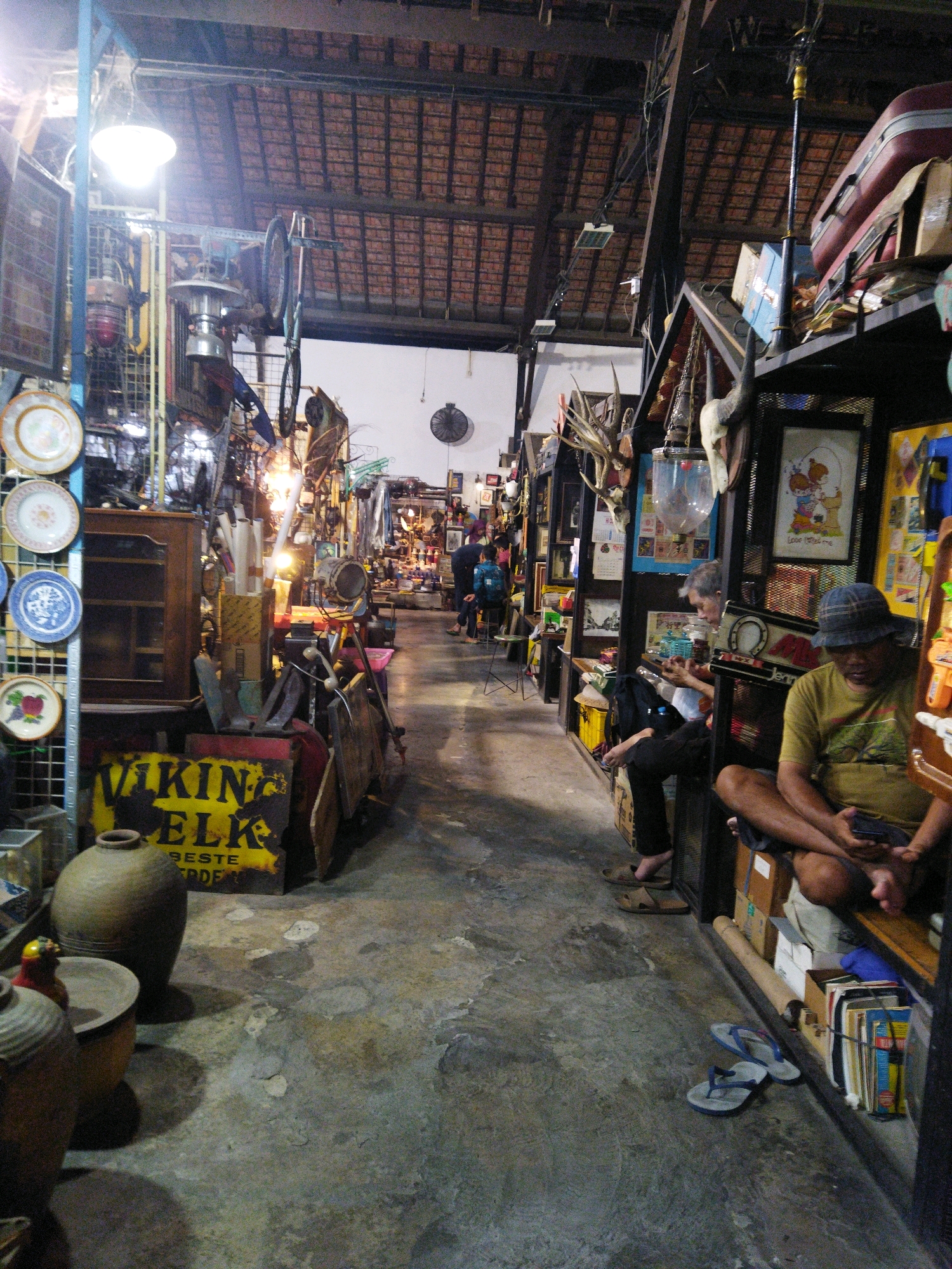 The image depicts the interior of a cluttered shop, possibly an antique or vintage store, located in Semarang, Indonesia. The space is narrow and filled with a diverse assortment of items, including decorative plates, lamps, wooden furniture, bicycles, and various trinkets. The floor is concrete and shows signs of wear.
In the foreground on the left, two large ceramic pots are visible. Behind them, a yellow sign with black lettering reads "VIKING ELK BESTE TRADE". To the right, a man wearing a hat and a green t-shirt is seated, looking at his phone. Another man is seated further back, also appearing to be engaged with his phone. In the background, several other individuals are present, some browsing and others walking through the aisle. The lighting is dim, with a mix of overhead lamps and natural light filtering in. The ceiling is made of wooden planks. The overall impression is of a densely packed, eclectic retail environment.
