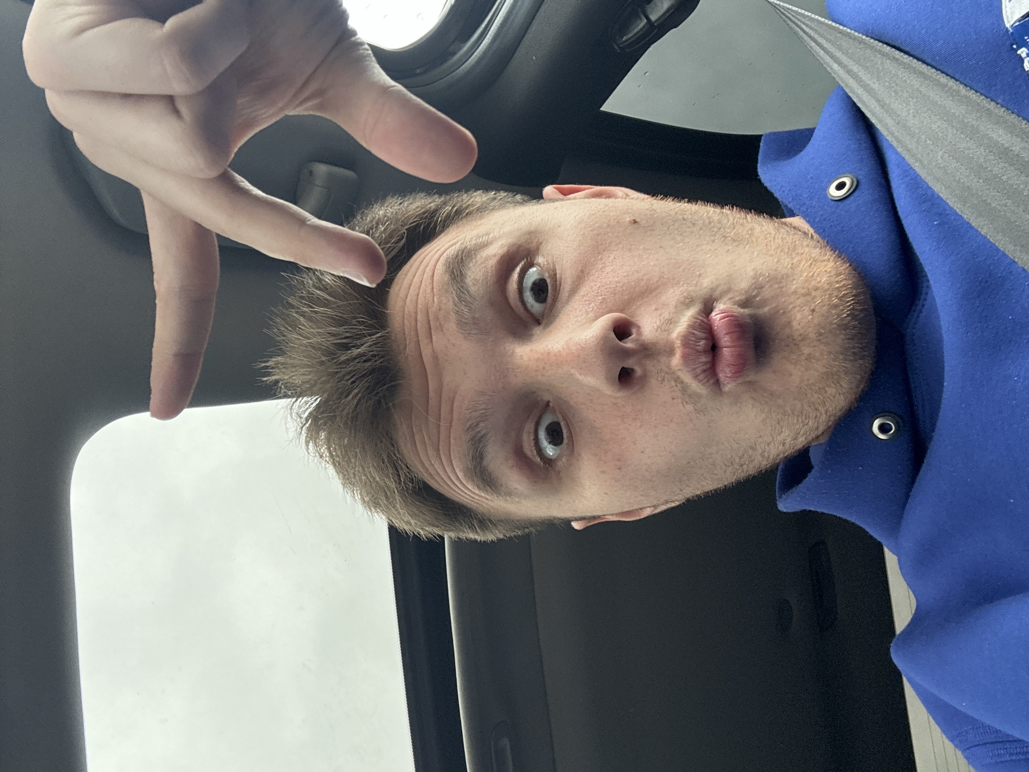 A man is inside a vehicle, wearing a blue hooded sweatshirt and a seatbelt. He is making a hand gesture with his fingers. The scene is inside of a car with a window showing an overcast sky. The location is undetermined and unknown.