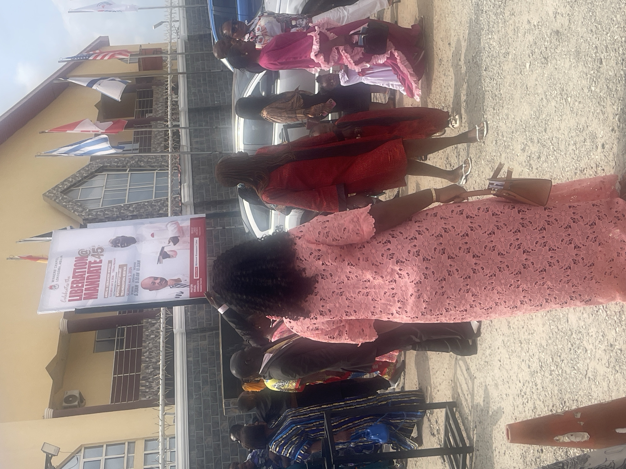 The image shows a group of people gathered outside Winners Chapel Lokogoma, Abuja, Nigeria. A banner promoting a "Liberation Mandate 45" event is visible. Flags, including what appear to be the American and Israeli flags, are displayed on poles in front of the building. Several individuals are dressed in formal attire, with one woman in the foreground wearing a pink lace dress. The surrounding environment appears to be a paved area.