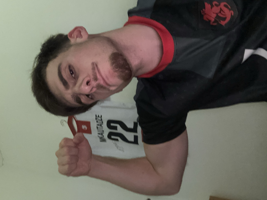 A fair-skinned man flexes, dressed in a gray and black jersey with a red dragon emblem and red trim. Behind him hangs a white basketball jersey labeled "MKAUTADZE 22". His expression is playful, with a slight smirk. The background consists of a light-colored wall, suggesting an indoor setting. The image is rotated slightly, adding a casual, selfie-style aesthetic.