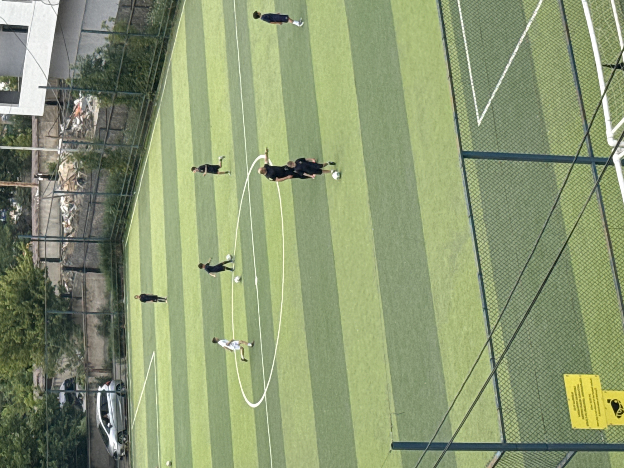This aerial photograph captures a group of individuals engaged in a soccer match on an outdoor artificial turf field. The field is marked with white lines, delineating the playing area. The setting appears to be a sports complex or park, with a building and lush green trees visible in the background to the left. A chain-link fence encloses the field.
Several people are scattered across the field, some actively playing with a soccer ball, while others are positioned as if participating in a practice or casual game. One individual in the center is in motion, seemingly about to kick or having just kicked the ball. Others are running or standing, observing the game. The players are dressed in athletic attire, primarily dark colored shirts and shorts, with one player in a white uniform.
The lighting suggests it is daytime, likely late morning or early afternoon, with bright sunlight casting shadows on the field. The weather appears to be fair and sunny, conducive to outdoor activities. The presence of a car parked outside the fenced area and the urban-like greenery hints at a location within a city.
On the fence at the lower right, two yellow signs are visible. One sign is primarily text-based, with an image of a person in silhouette holding a ball above their head, possibly indicating a warning or instruction related to the facility. The second sign has more text, but it is largely illegible in this resolution. The overall impression is a vibrant sports scene on a clear day in an urban environment.