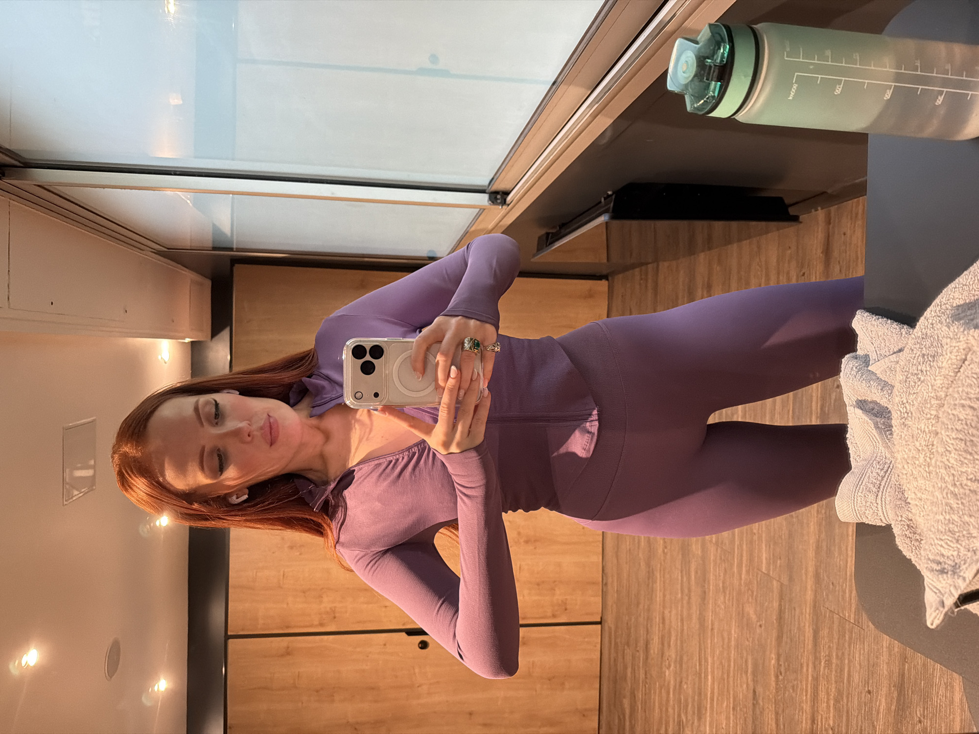 A woman with red hair is taking a selfie in what appears to be a locker room or changing area. She is wearing a purple athletic outfit consisting of a long-sleeved top and leggings. She holds a smartphone in front of her, capturing her reflection in a mirror.
Behind the woman, there is a window with a partial view of the outside. A pale green water bottle is situated on a surface near the mirror. A white towel rests on the edge of a counter or bench.
The walls and cabinets in the room are constructed of wood. The scene is lit with a warm light, possibly from overhead fixtures. The location is not identifiable as Paris, France, based on the provided image.