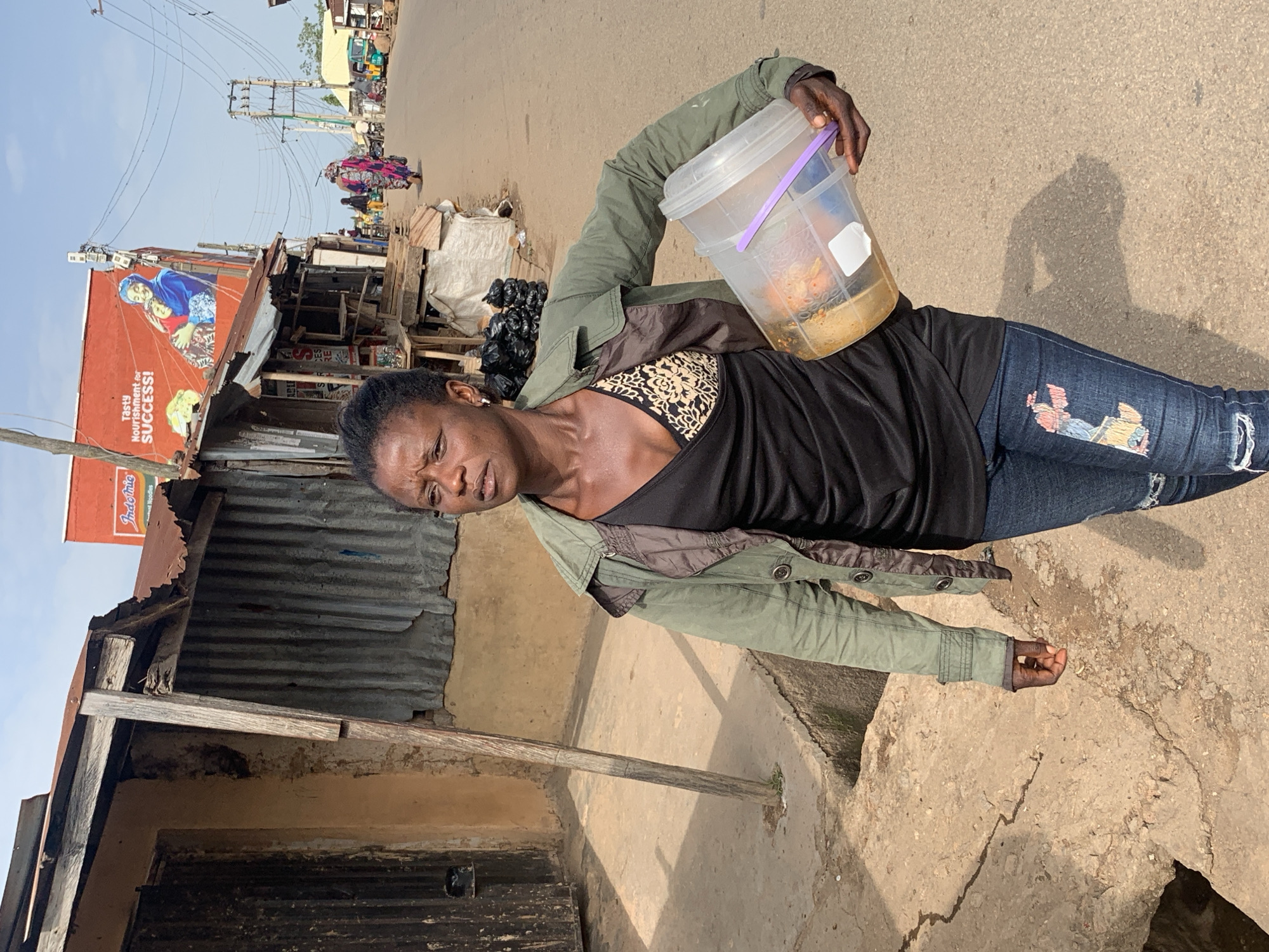 In Jos, Nigeria, a woman walks down a dusty street, her face etched with weariness. She carries a large, clear plastic bucket filled with food, its purple handle gripped firmly in her hand. The bucket appears to be made of recycled materials, common in local markets. She wears a green jacket over a black top with a leopard print bra, and ripped blue jeans. The background reveals a bustling street scene, with dilapidated buildings constructed from corrugated metal and mud. A large, colorful billboard advertises "Indomie Noodles" with the slogan "Tasty Nourishment for SUCCESS!". People are seen walking and riding motorcycles in the distance, with more stalls and makeshift shops lining the road, typical of a market environment in Jos. The overall impression is one of resilience amidst hardship, a common theme in urban settings in Nigeria.
