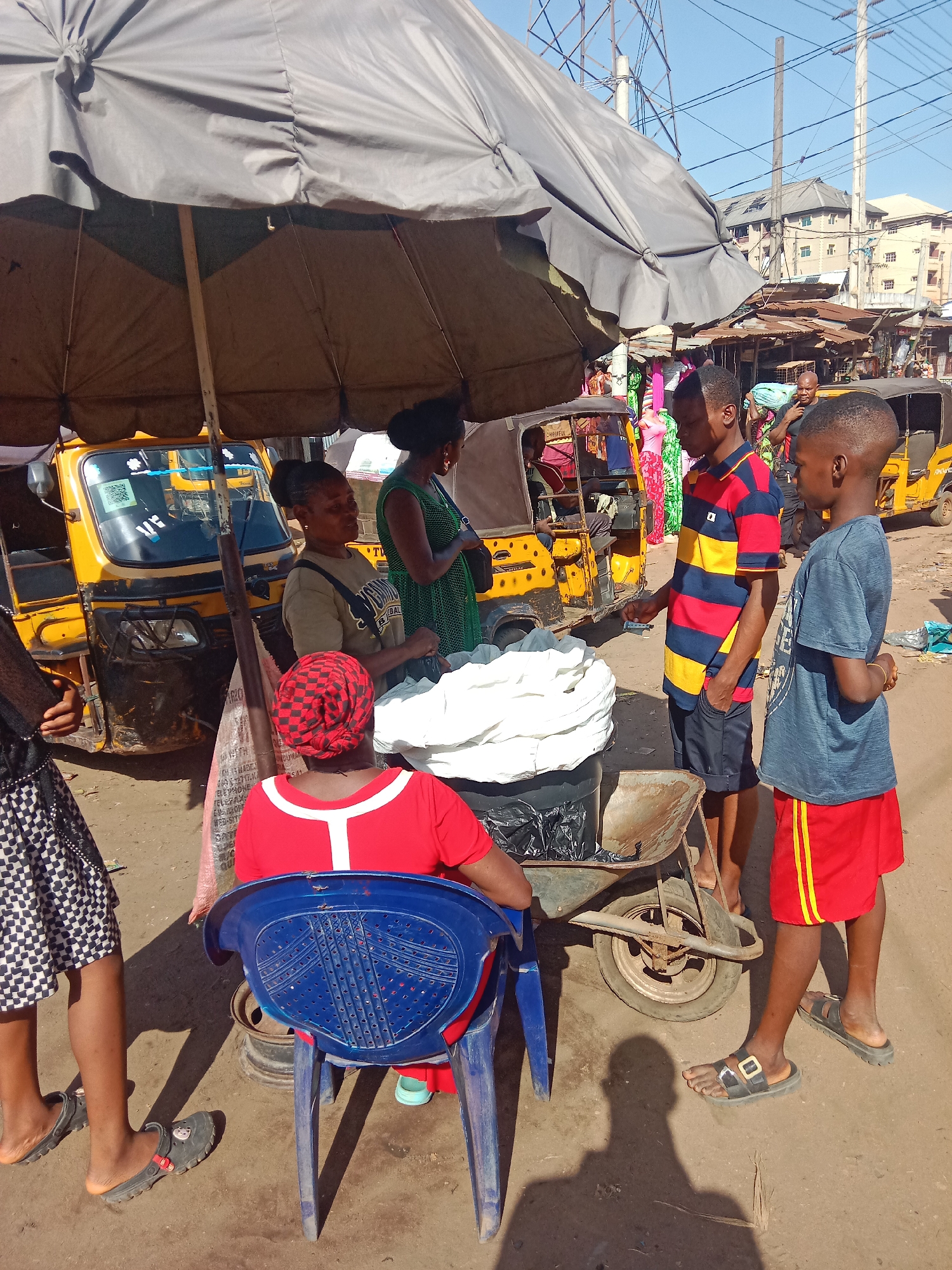 This image captures a vibrant outdoor market scene in Nkpor, Nigeria, under a bright, sunny sky, likely midday given the strong shadows. A large grey market umbrella provides shade over a makeshift stall.
At the center, a woman in a red top and red-and-black checkered headwrap is seated on a blue plastic chair, her back to the viewer. She is positioned in front of a stall made partly from a rusty wheelbarrow, which holds a substantial pile of white, possibly food, items. Standing behind the stall and conversing with customers is another adult woman wearing an olive-green shirt and a dark shoulder bag. Further behind her, a woman in a green patterned dress is visible.
Two boys are engaging with the stall. One boy, wearing a red, yellow, and blue striped polo shirt and dark shorts, stands facing the stall with what appears to be money in his hand. Beside him, another boy in a blue t-shirt and red shorts with yellow stripes stands observing. To the far left, the lower half of a person wearing a checkered skirt and dark slippers is partially visible.
In the background, several yellow tricycle taxis, commonly known as Keke Napep, are parked, indicating a busy commercial area. The text "CHEERFUL" is visible on the front of one Keke. The background also shows multi-story buildings, dense power lines, and other market stalls, reinforcing the urban market setting. A sign partially visible behind the blue chair includes words like "TELEFAX" and "WEBSITE." The scene depicts a typical daily market interaction in a bustling Nigerian street.