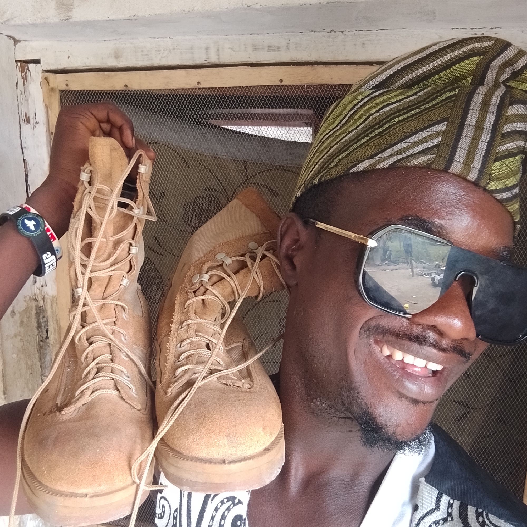 In the city of Ado, Nigeria, a person is holding up two tan combat boots. They are wearing a patterned headwrap and sunglasses, and are smiling. The background consists of a door with a mesh screen and a wall. A wristband is visible on the person's left wrist.
