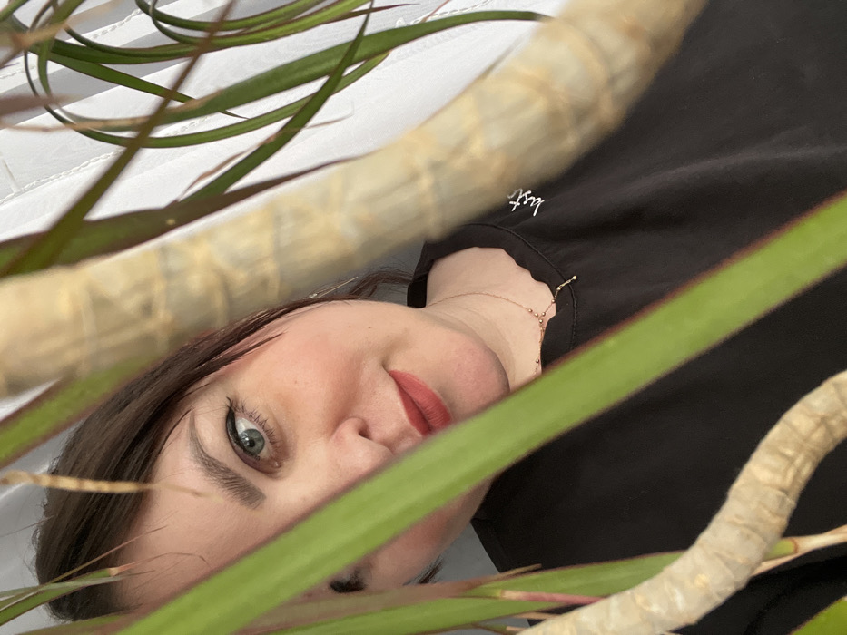 The image captures an indoor scene in Noviy, Russia, featuring an adult female partially obscured by a houseplant. The subject's right eye, nose, mouth, and chin are visible, framed by dark hair. She has blue-green eyes, wears red lipstick, and displays a subtle smile, looking directly towards the viewer. She is attired in a dark, likely black, crew-neck top with white embroidery on the right chest area, which appears to spell "just". A gold-colored necklace with small spherical beads and a linear dangling element is visible around her neck. The foreground is dominated by the light brown, textured stems and long, slender green leaves of a plant, consistent with a Dracaena species, which partially block the subject's left facial features and upper body. The background consists of a light-colored, vertically patterned surface, suggesting a wall or window blinds.