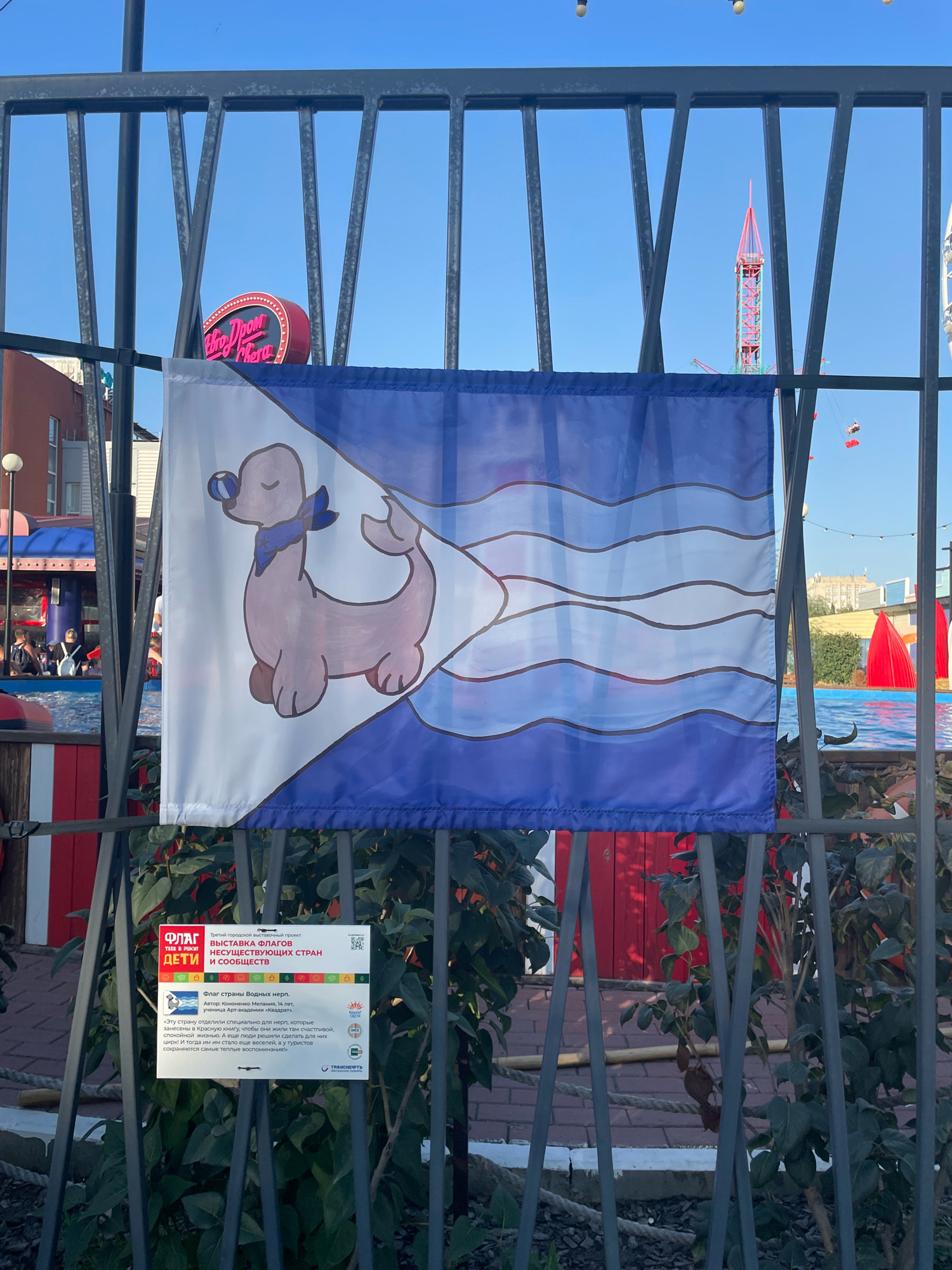 The image shows a flag with a cartoon seal on it, displayed on a black metal fence. The flag is white on the left and blue on the right, with wavy blue lines on the blue portion. The seal is pinkish-brown with a blue scarf and a blue ball in its mouth.
Below the flag, a sign with Russian text is attached to the fence. The sign indicates it is part of a project called "Flags of Countries" for children. The title of the exhibit is "Flags of Countries of Unusual Nations and Societies". The text mentions a country of "Water Seals" and describes how they are kept in a red book.
In the background, there is a body of water, likely a pool, with red poolside structures. Further in the background, there are buildings, a Ferris wheel, and what appears to be an amusement park ride. The sky is clear and blue.
The sign details:
The project is titled "Flags for Children."
The specific exhibit is "Flags of Countries of Unusual Nations and Societies."
The author of the flag is Kononenko Melani, 14 years old, a student of the Art Academy "Kvadrata."
The flag represents the "Country of Water Seals."
The overall scene suggests an outdoor event or exhibition in an amusement park or recreational area. Based on the provided information, the location is the city of Omsk, Russia.