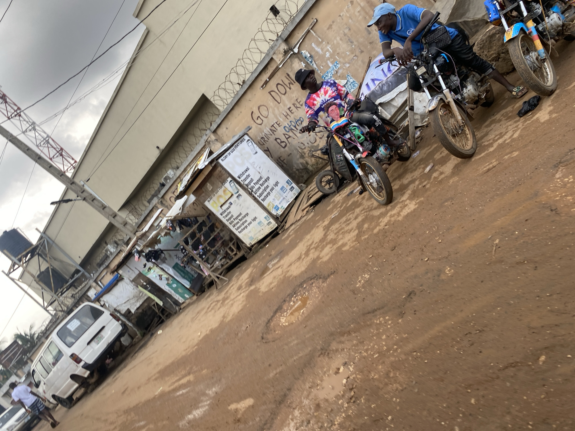 The image shows a street scene in Lagos, Nigeria. Two men are sitting on motorcycles on a muddy, uneven road. One man wears a blue shirt and cap, while the other wears a tie-dye shirt and a bucket hat. The motorcycles are of different colors and designs. Behind them is a weathered building with "GO DOW URINATE HERE Y BARP" spray-painted on the wall. Barbed wire runs along the top of the wall. A small roadside business with signs advertising financial services, including "GLORY OF GOD VENTURES", stands to the left. A white van is parked near the roadside business. A telecommunications tower is visible in the background against a cloudy sky. Puddles and debris are scattered on the ground.