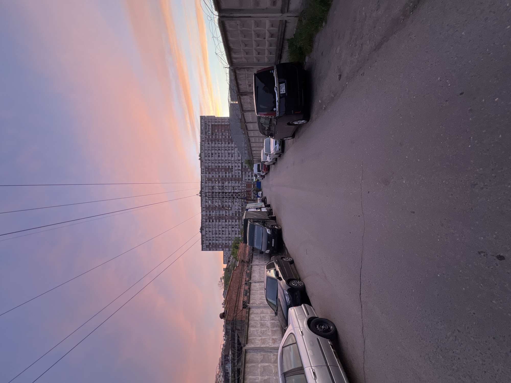 The image shows a street in what appears to be an urban area with a row of parked cars along the side of a concrete wall. A tall, multi-story building is visible in the background, possibly a residential apartment complex. The sky is filled with clouds at sunset. Overhead power lines are seen across the sky. Based on the car license plates, the location is Krasnodar, Russia.